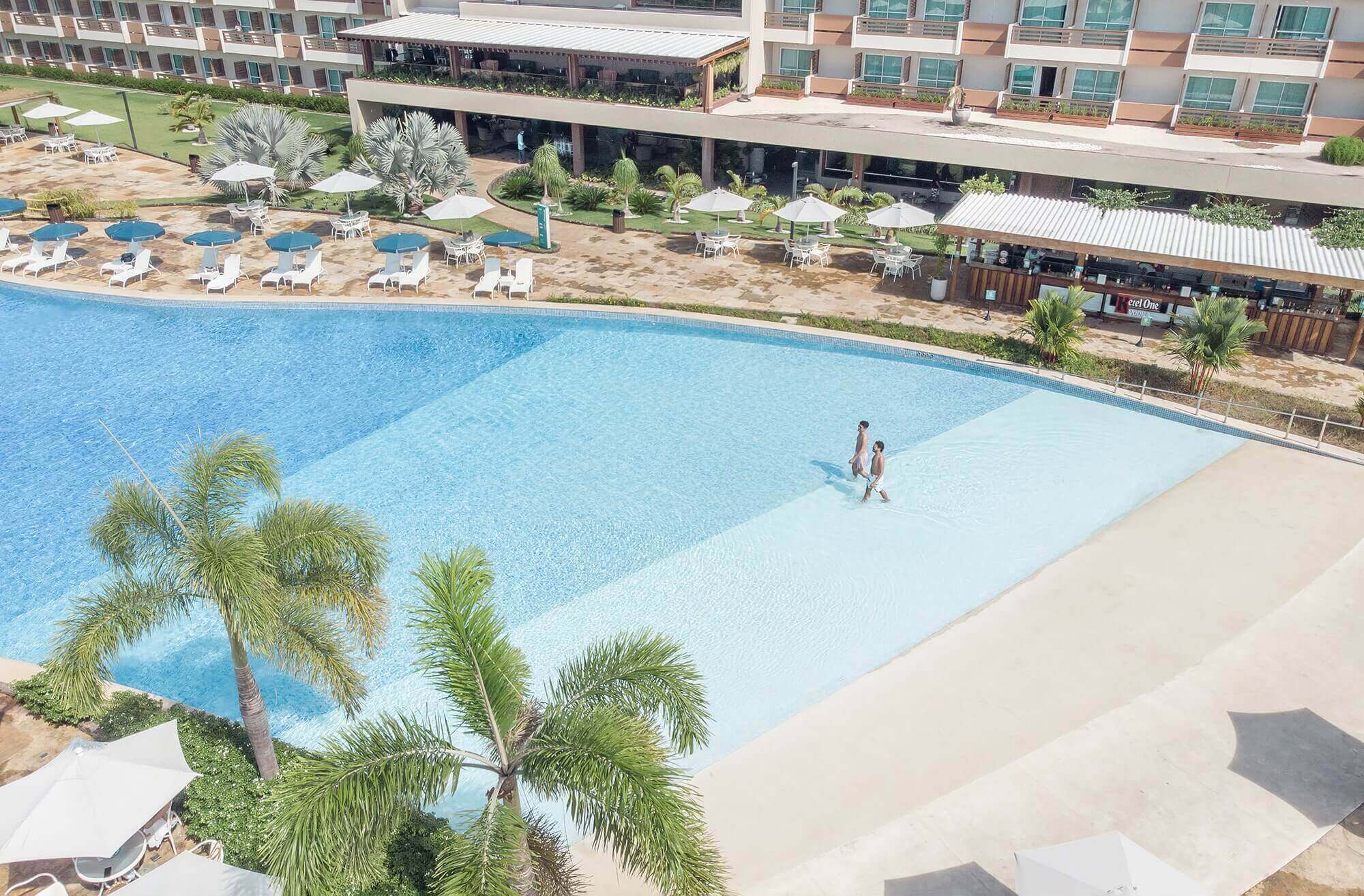 Vista aérea de dois homens entrando na piscina. Guarda-sóis, espreguiçadeiras e estrutura do resort ao fundo.