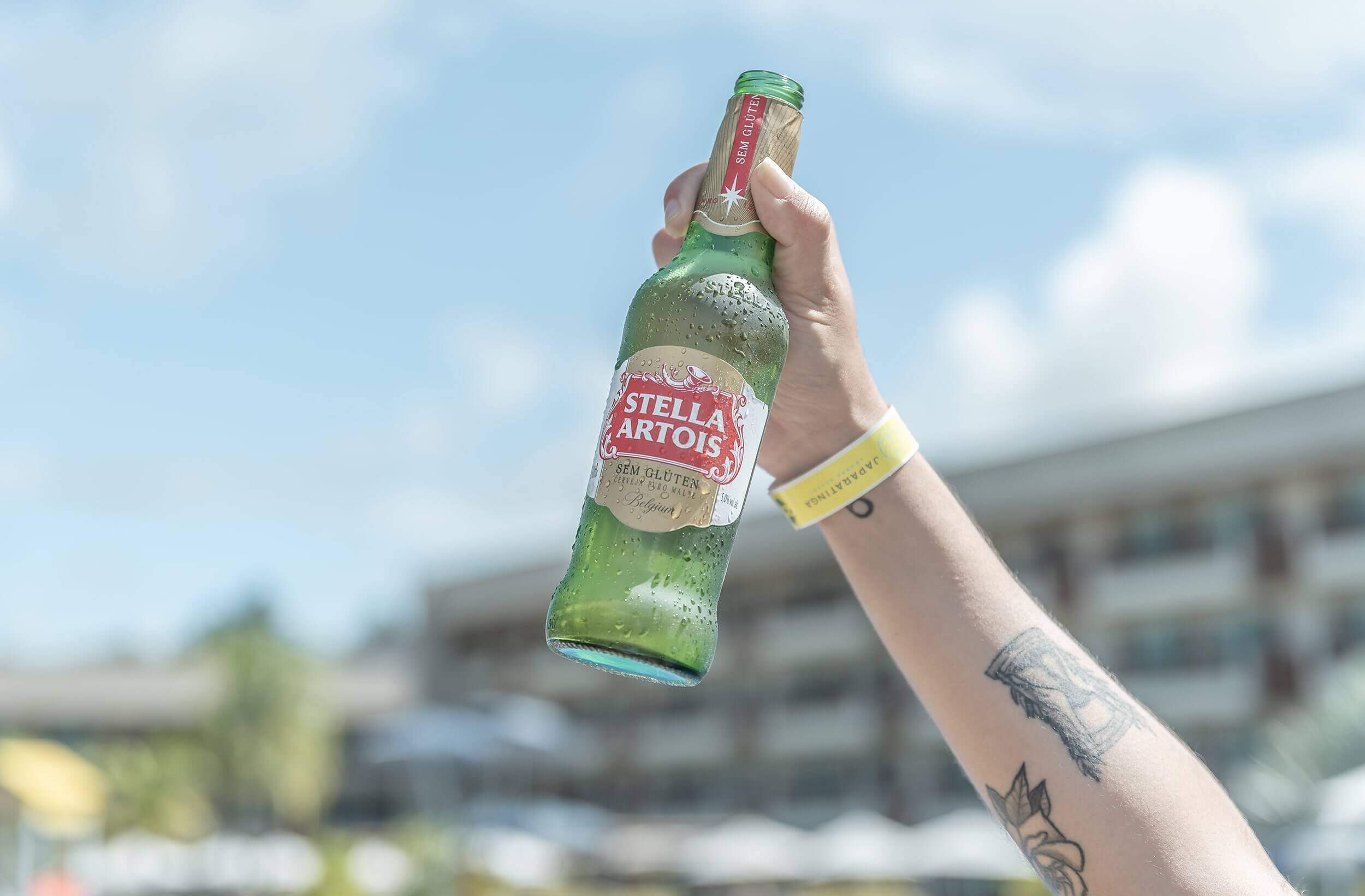 Mão segurando garrafa longneck de cerveja Stella Artois no alto, fundo desfocado.