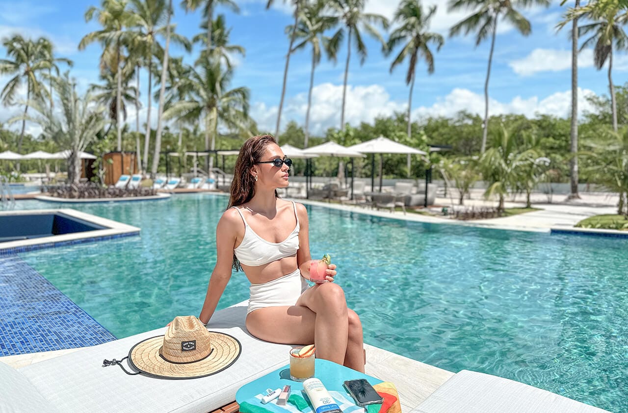 Na imagem, há uma mulher sentada na borda de uma piscina infinita. Ela está vestindo um biquíni branco e óculos escuros, e segura uma bebida tropical, possivelmente um coquetel, com um enfeite de fruta. Ao lado dela, sobre uma toalha de praia colorida, estão um chapéu de palha, protetor solar, um telefone celular e outros itens pessoais.