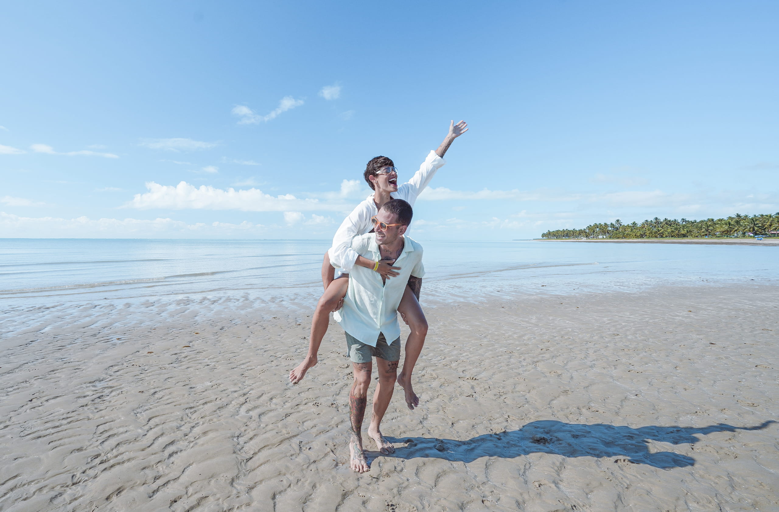 Vista de um casal caminhando sobre uma grande faixa de areia que emerge em águas rasas e cristalinas. A mulher está pendurada nas costas do homem enquanto ambos sorriem, com o horizonte distante de mar, vegetação e céu.