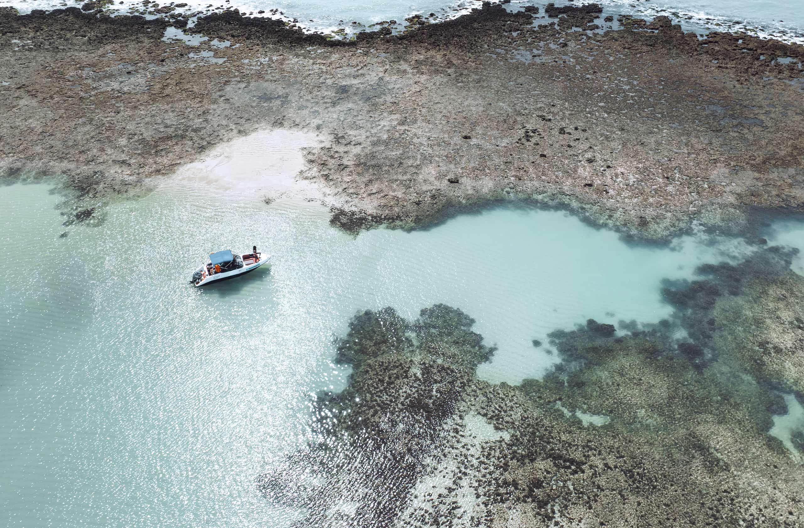 Vista aérea de cima de extensos recifes de coral e piscinas naturais na praia de Japaratinga. As águas são azul-turquesa e verde-claras, com ondas quebrando na barreira de recifes. Um pequeno barco e pessoas são visíveis na área rasa e calma.