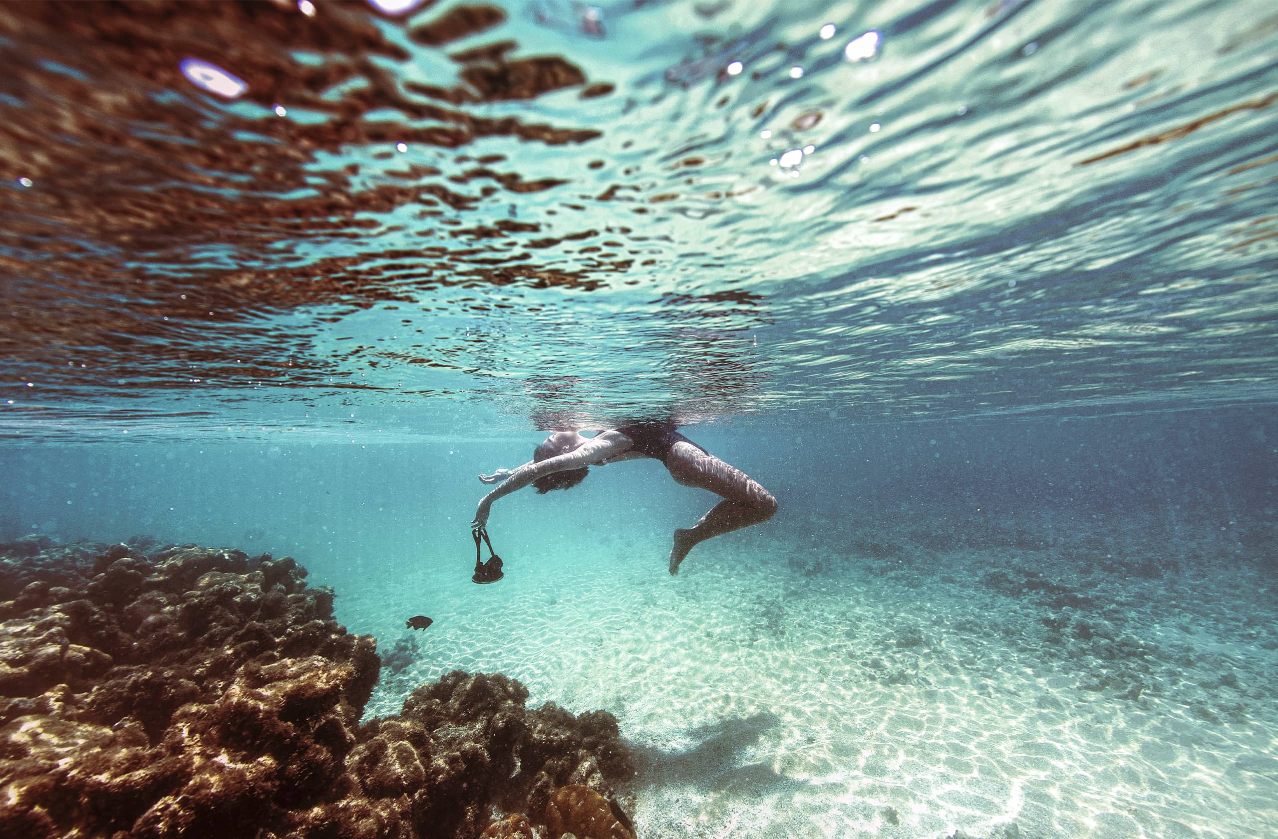 Foto subaquática de uma pessoa flutuando na superfície da água, perto de um recife de coral. A água é cristalina, com o fundo de areia visível, e a luz do sol cria ondulações na superfície.