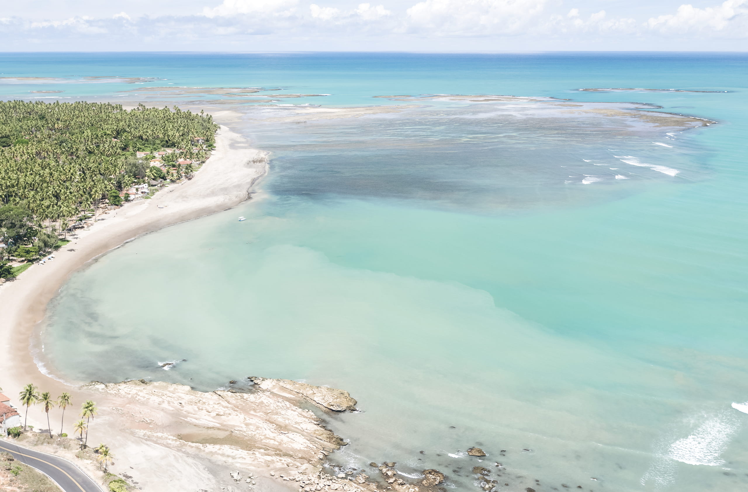 Praia de Japaratinga vista de cima, com uma área rochosa que se conecta a uma faixa de areia que forma uma curva. O oceano exibe tons de azul e verde, com extensos recifes de coral visíveis na maré baixa. A costa é ladeada por coqueiros e vegetação.
