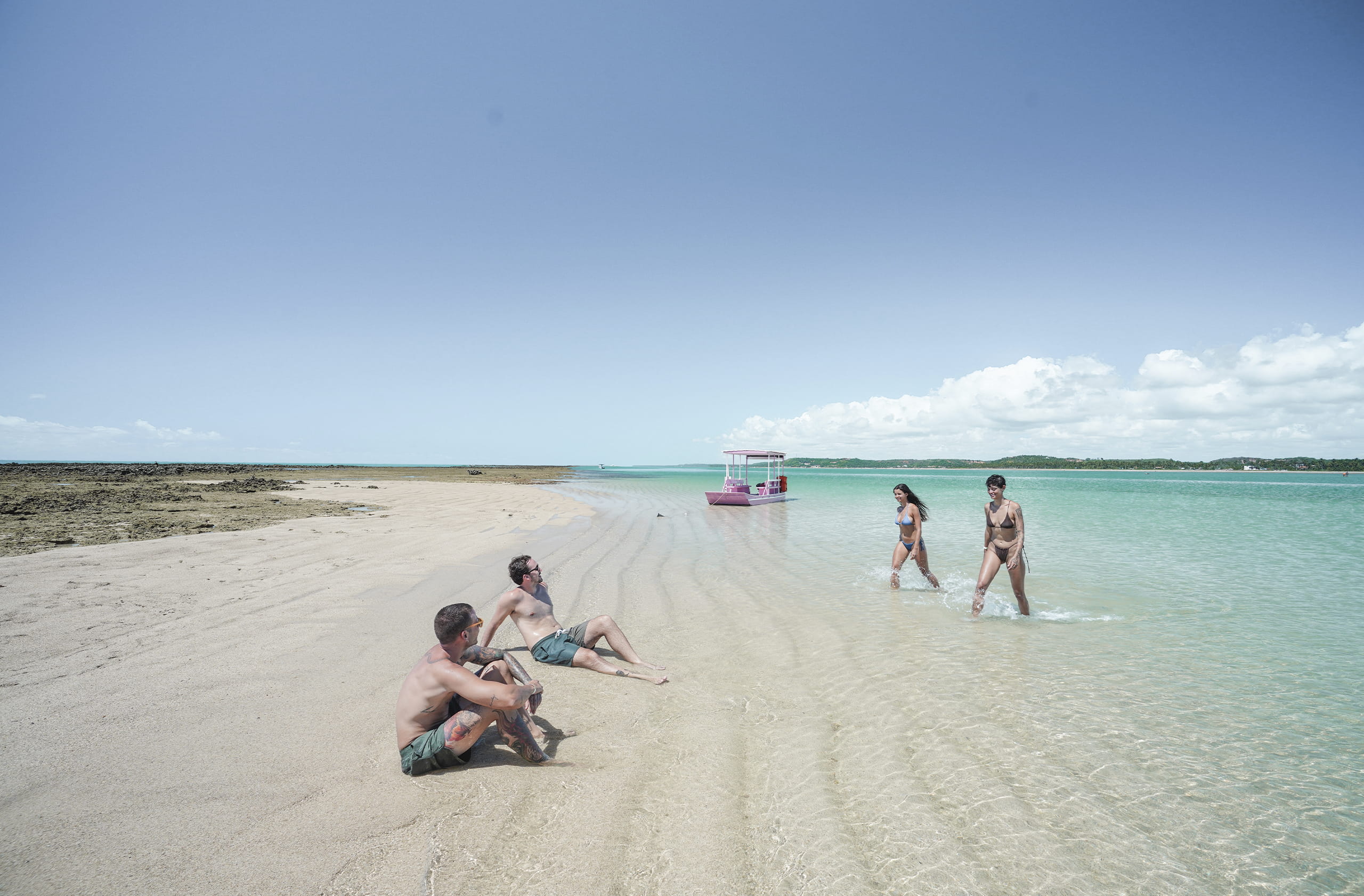 Grupo de amigos jovens na Croa de São Bento, enquanto as mulheres caminham em águas rasas e claras da praia, os homens estão sentados na faixa de areia. Ao fundo, uma imensidão do mar com um céu azul.