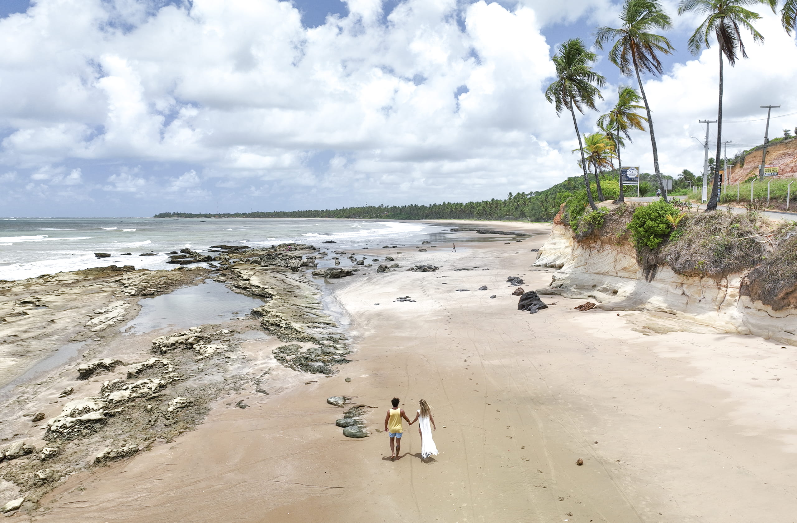 Casal caminhando descalço na areia da praia de Barreiras do Boqueirão à vista de cima. A areia tem marcas de ondulação e há rochas aparentes, e a água calma e rasa se estende ao fundo, coqueiros nas laterais da costa e céu com algumas nuvens.