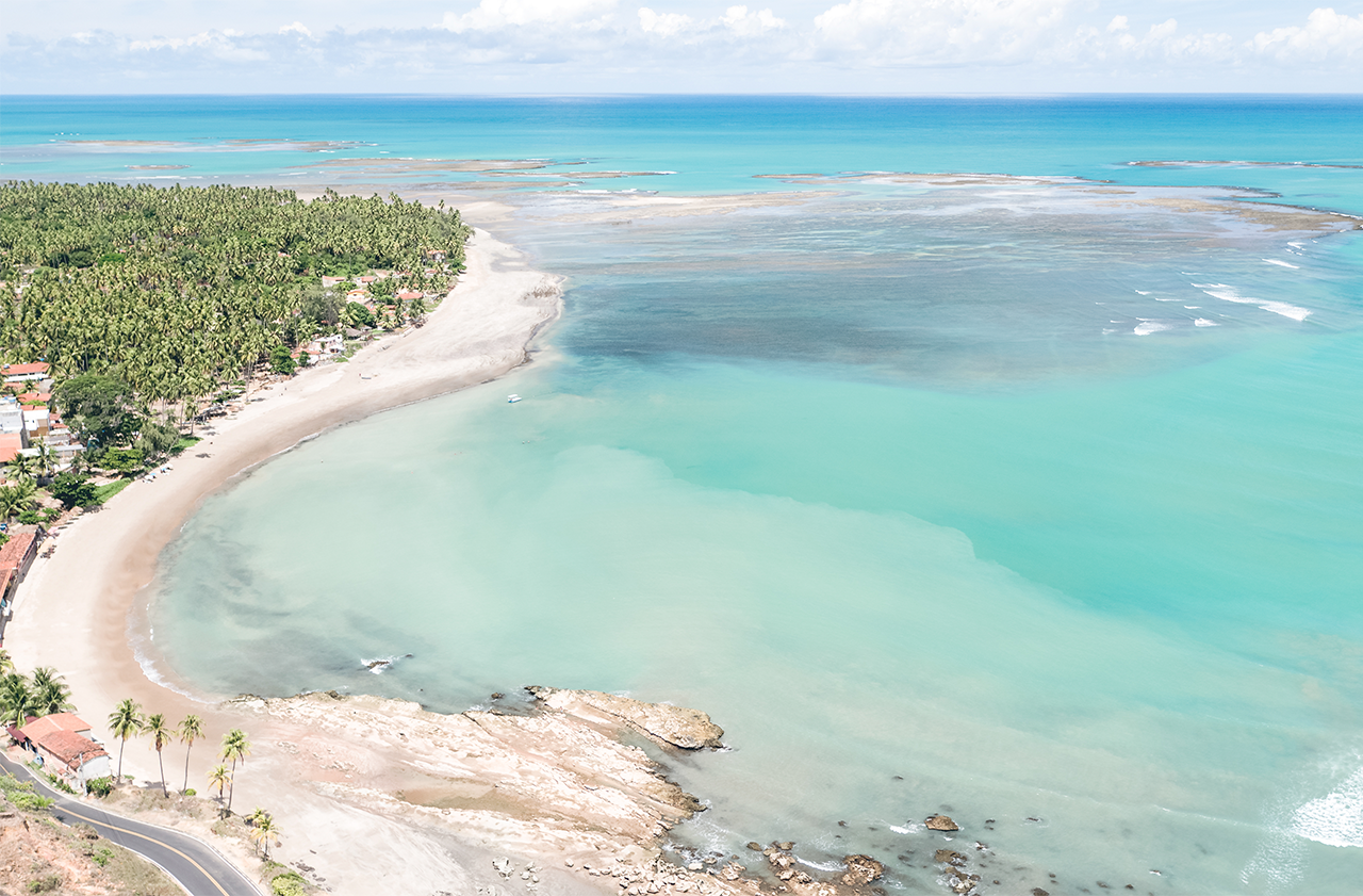 Praia de Japaratinga vista de cima, com uma área rochosa que se conecta a uma faixa de areia que forma uma curva. O oceano exibe tons de azul e verde, com extensos recifes de coral visíveis na maré baixa. A costa é ladeada por coqueiros e vegetação.