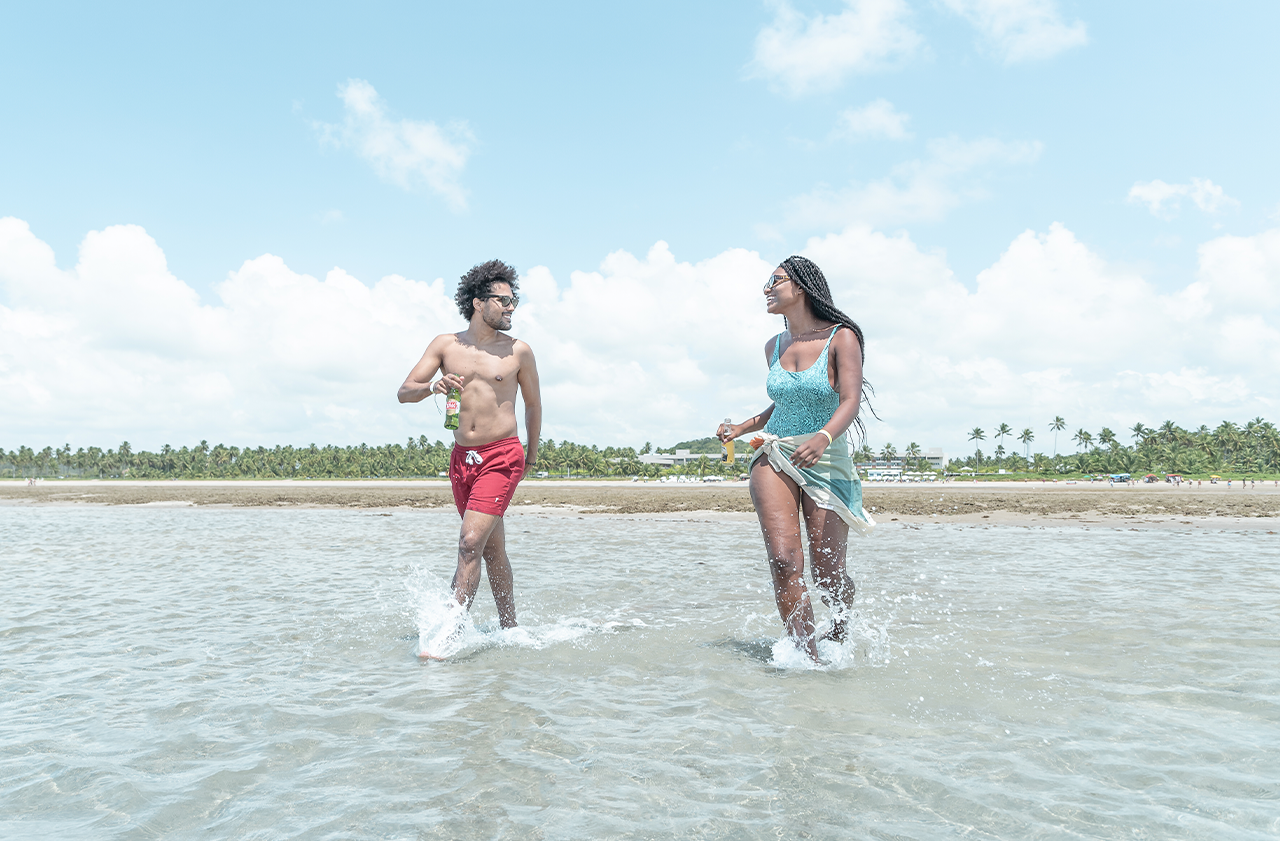 Um casal jovem, sorrindo e respingando água, caminha em águas rasas e claras da praia. Ao fundo, uma faixa de areia e coqueiros sob um céu azul com nuvens.