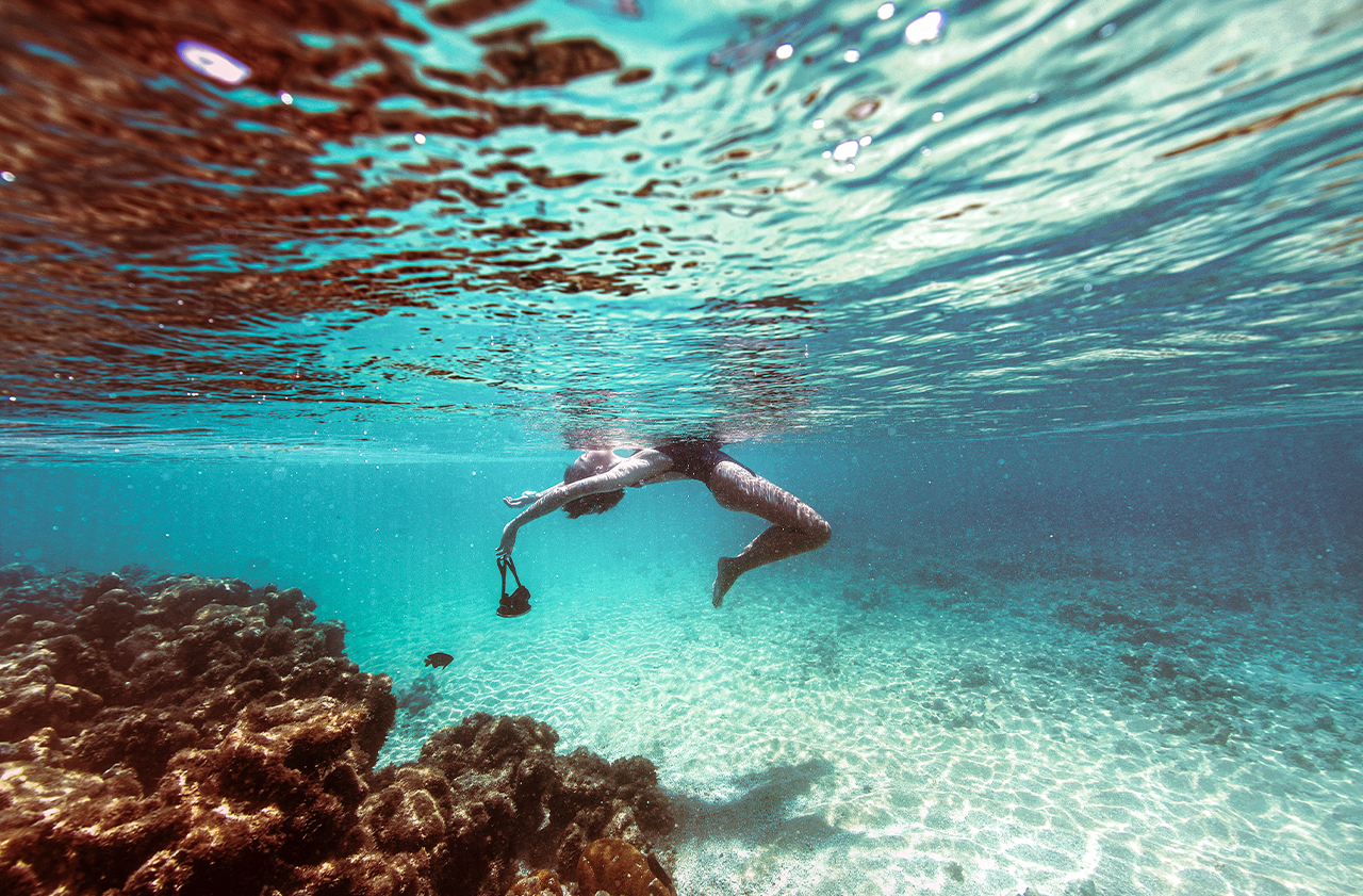 Foto subaquática de uma pessoa flutuando na superfície da água, perto de um recife de coral. A água é cristalina, com o fundo de areia visível, e a luz do sol cria ondulações na superfície.