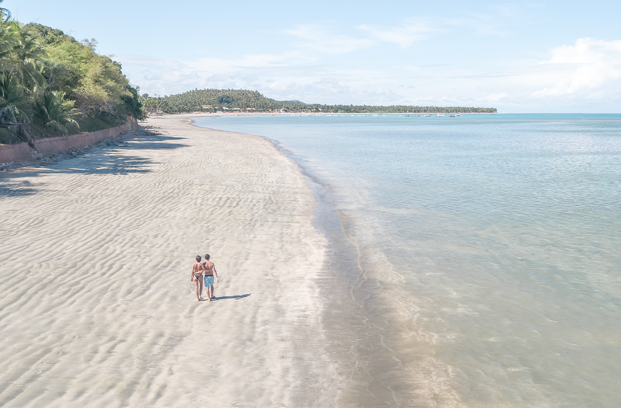 Casal caminhando descalço na areia da praia vista de cima. A areia tem marcas de ondulação, e a água calma se estende ao lado de uma costa com vegetação, coqueiros ao fundo e céu com algumas nuvens.