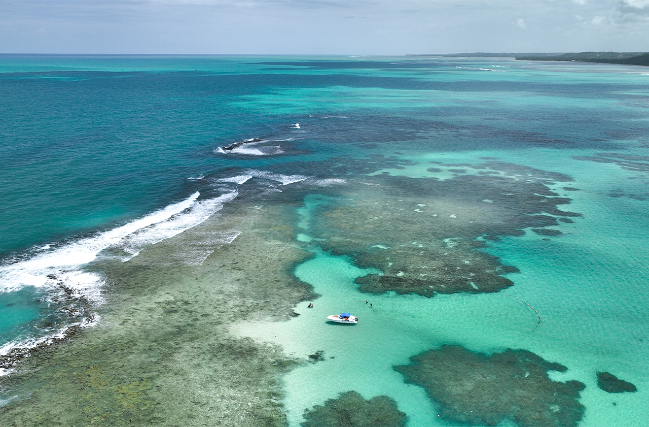Vista aérea de cima de extensos recifes de coral e piscinas naturais na praia de Japaratinga. As águas são azul-turquesa e verde-claras, com ondas quebrando na barreira de recifes. Um pequeno barco e pessoas são visíveis na área rasa e calma.