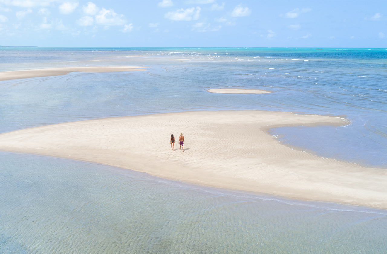 Vista de um casal caminhando sobre um grande banco de areia que emerge em águas rasas e cristalinas. A areia clara é cercada pelo oceano em tons de azul-claro, com o horizonte distante.