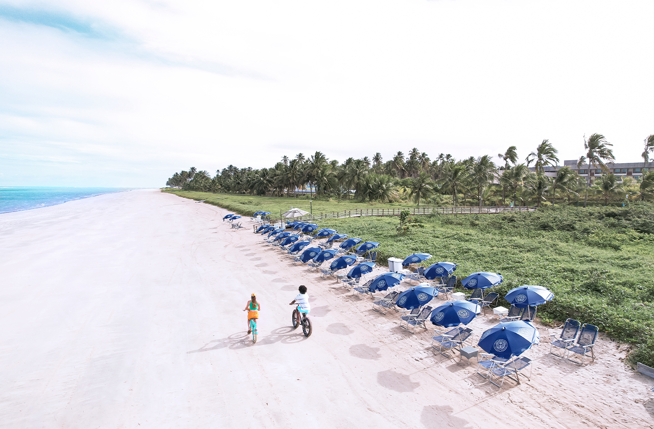 Praia larga e ensolarada vista de cima, com uma longa fileira de guarda-sóis azuis e cadeiras. Um casal anda de bicicleta na areia. Ao fundo, palmeiras e um edifício de resort moderno são visíveis.