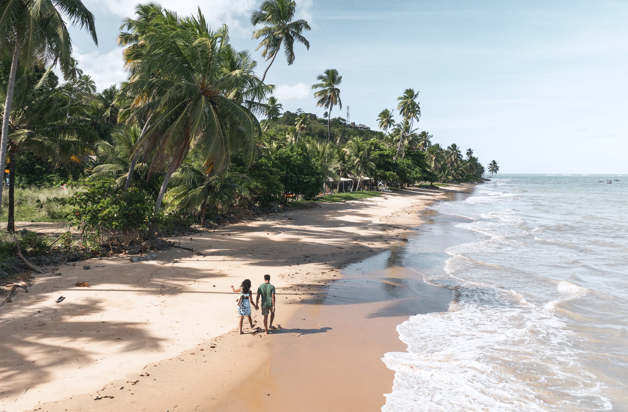 Casal caminhando descalços na Praia de Bitingui ensolarada. O sol forte brilha no alto do céu, refletindo na areia e no mar, que se estende ao horizonte com vegetação ao lado esquerdo.