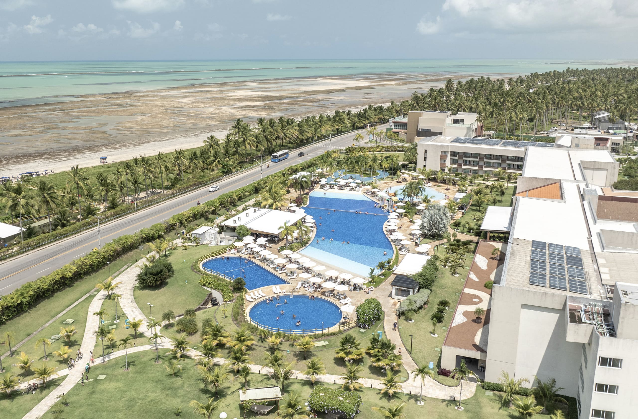 Vista aérea do Japaratinga Lounge Resort cercada por mata verde, e o no fundo a praia na maré baixa, mostrando vários recifes de coral e piscinas naturais onde pessoas estão andando na faixa de areia.