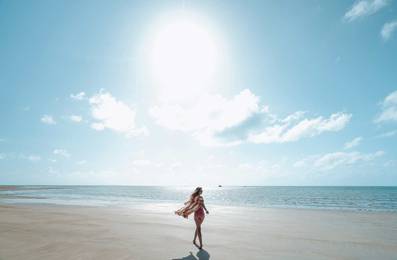 Uma mulher de biquíni e canga caminha descalça na praia ensolarada. O sol forte brilha no alto do céu, refletindo na areia e no mar, que se estende ao horizonte.
