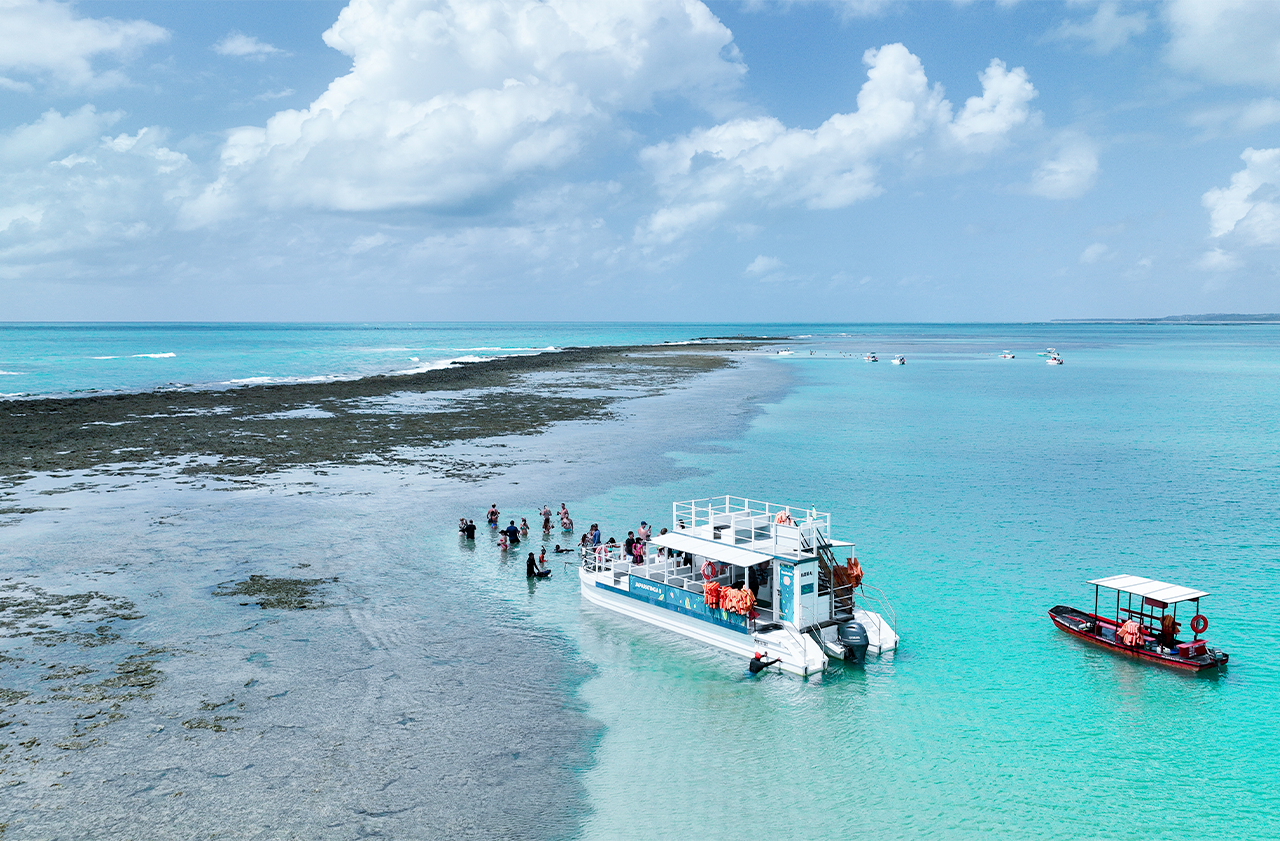 Vista aérea de um catamarã e um pequeno barco em águas rasas e cristalinas. Ao lado, um extenso recife de coral está exposto, com várias pessoas nadando e caminhando nas piscinas naturais formadas. O céu é azul com nuvens brancas.