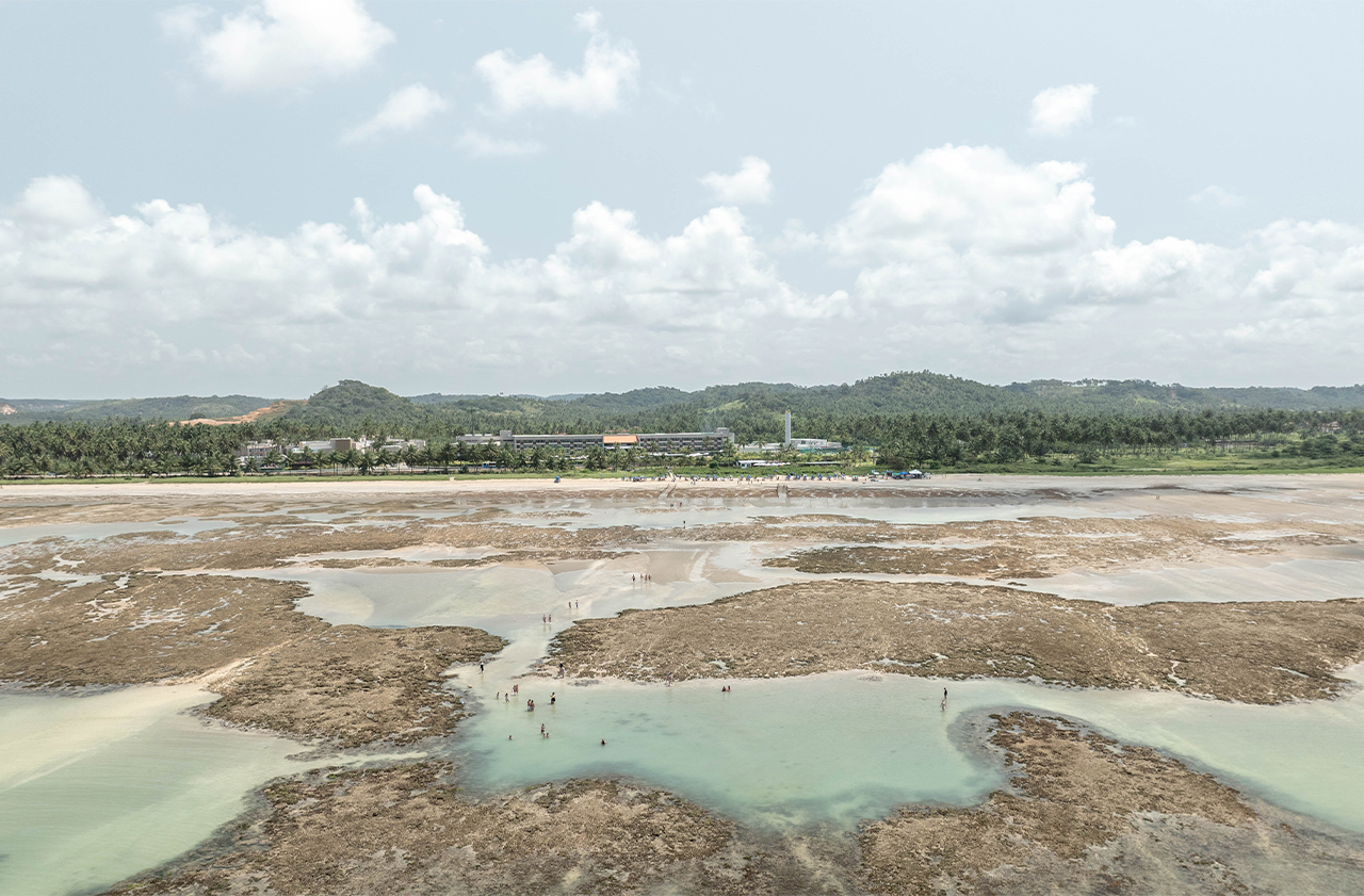 Vista da praia na maré baixa, mostrando vários recifes de coral e piscinas naturais onde pessoas estão andando. Na costa, uma faixa de areia larga é cercada por mata verde e o Japaratinga Lounge Resort no fundo.
