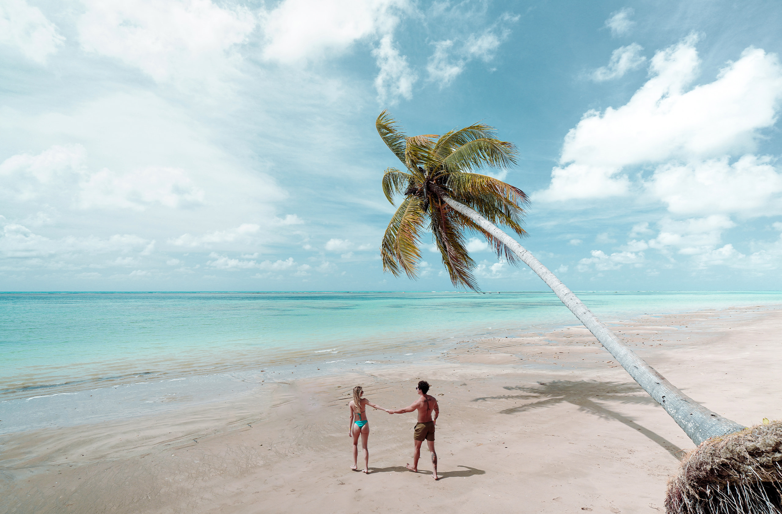 Casal de mãos dadas caminhando na praia, com o mar azul claro ao fundo e alguns barcos na água. Palmeiras e outras plantas na areia da praia.