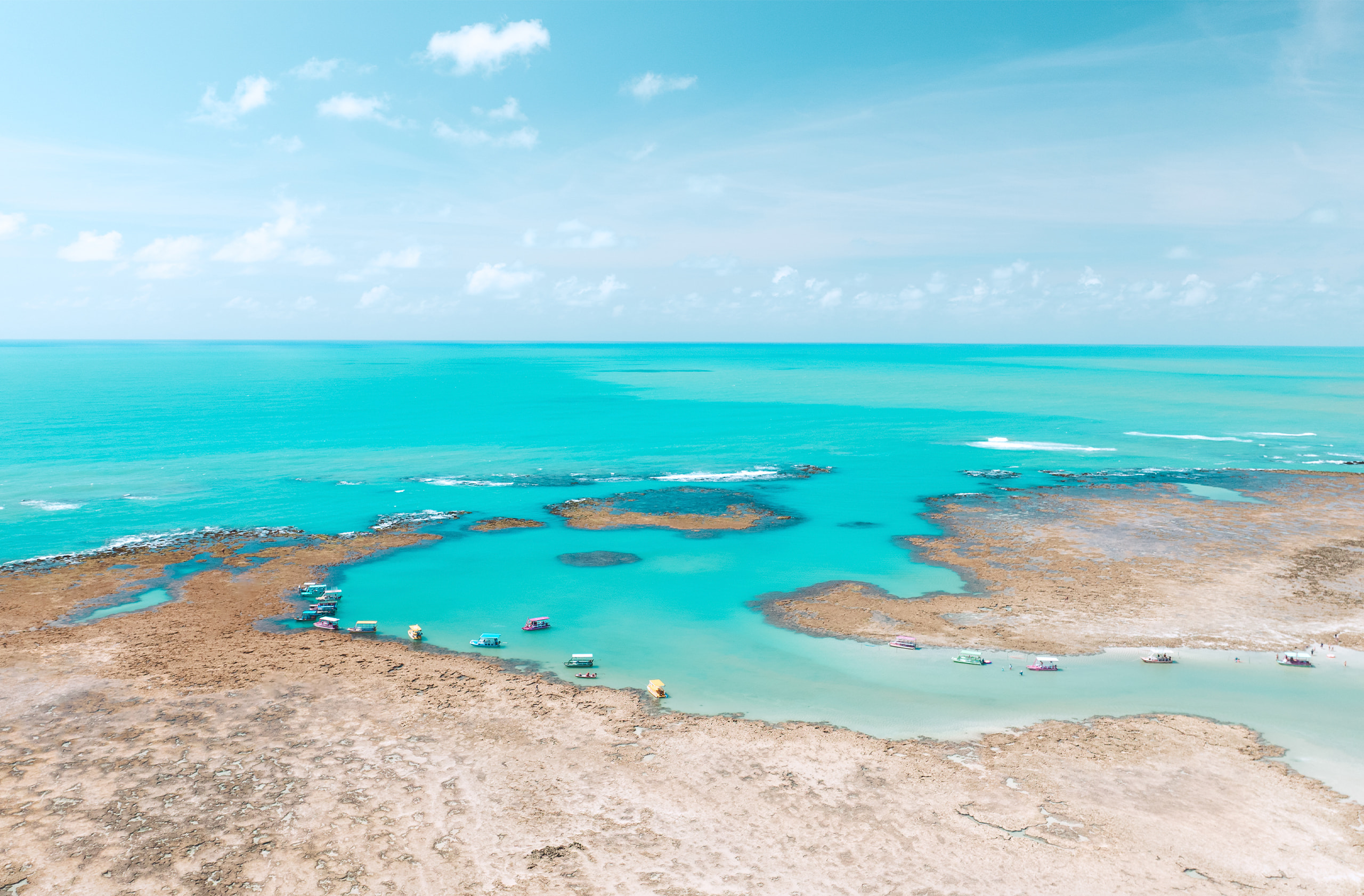 Vista aérea das piscinas naturais em um mar de águas cristalinas, formadas por recifes de corais. Pequenos barcos estão ancorados nos arredores.