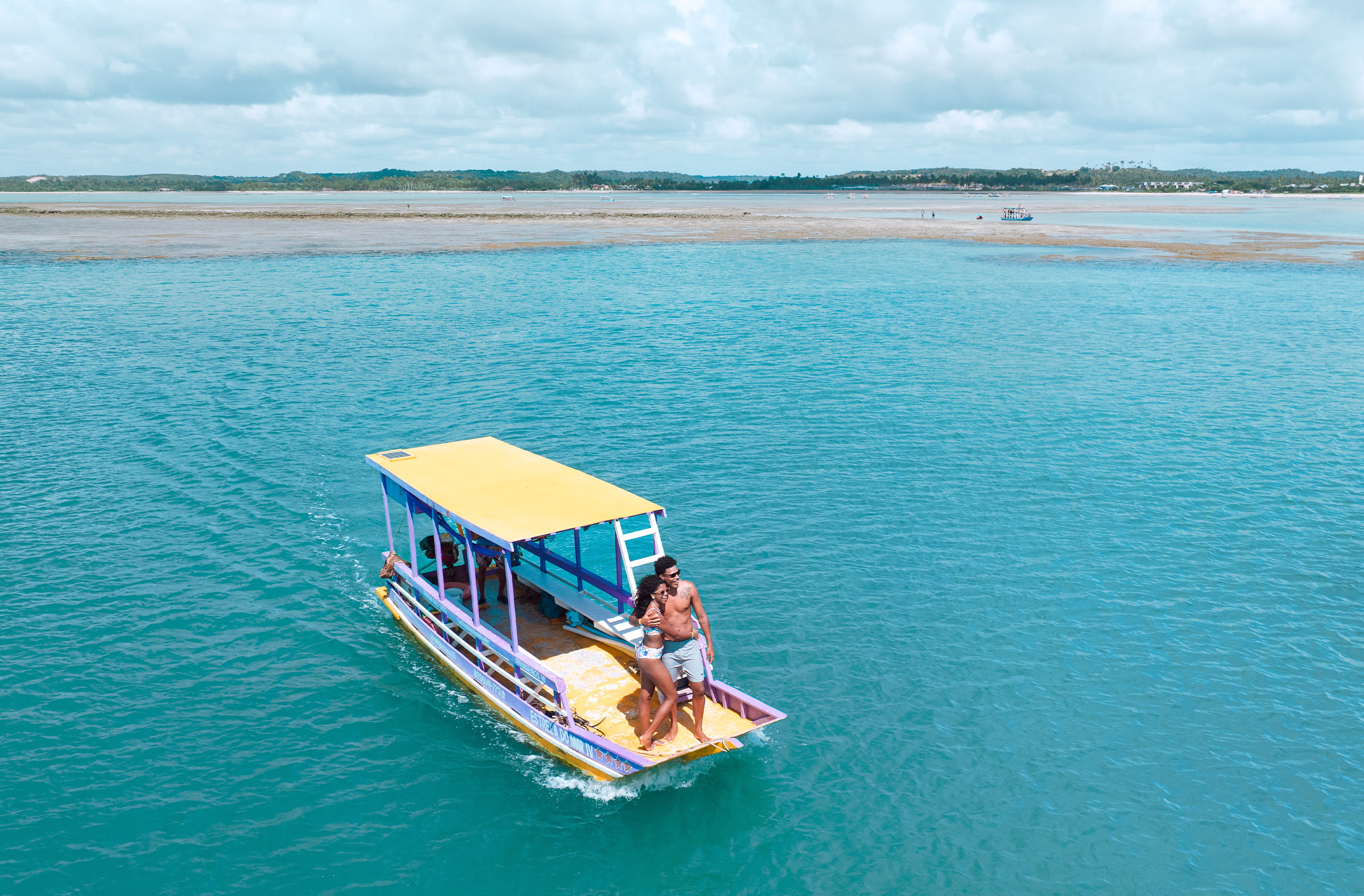 Vista de cima da praia com águas claras e calmas. Vários barcos pequenos e coloridos estão ancorados na água, e a costa tem areia branca e muitas palmeiras.