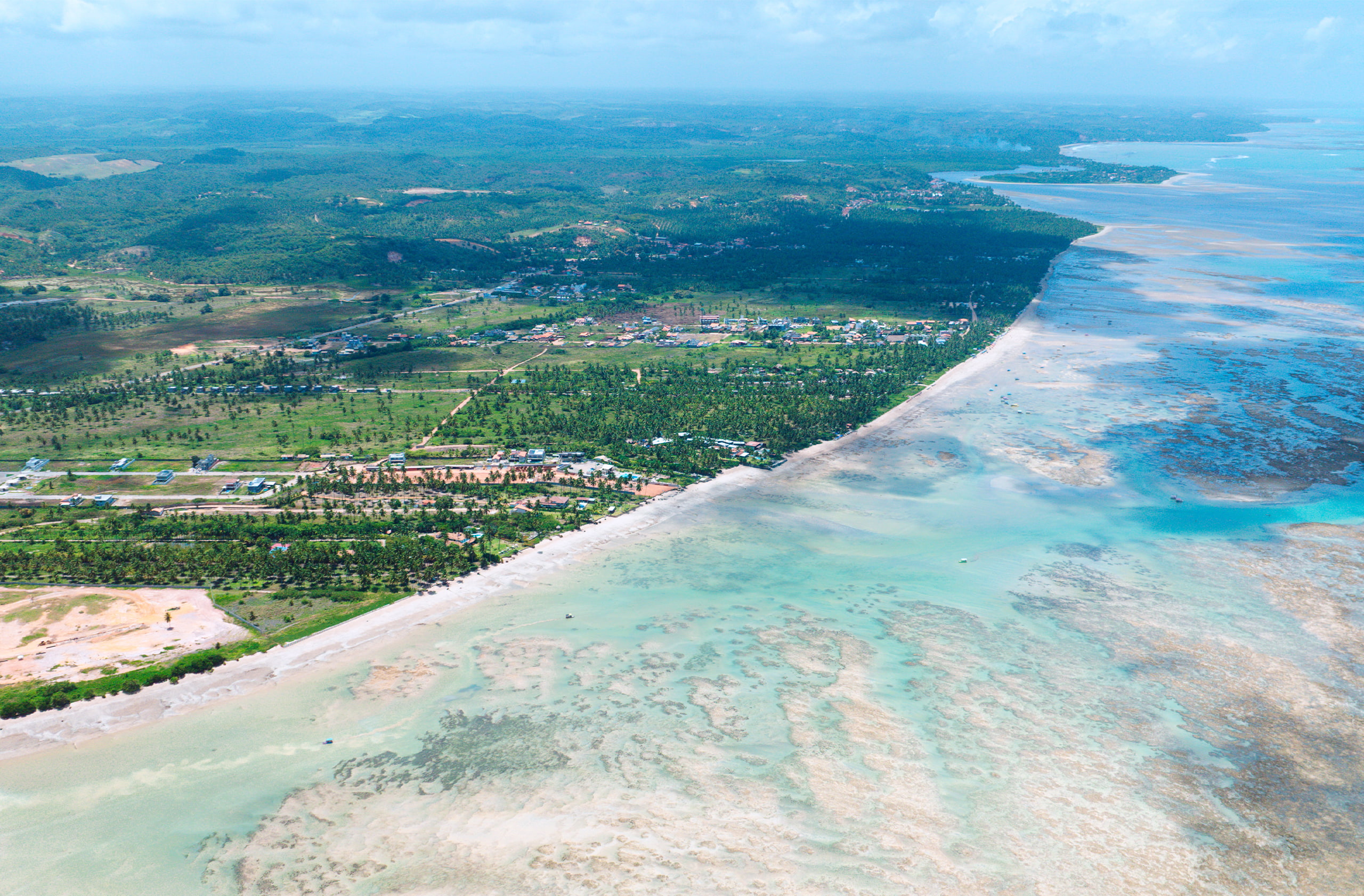 Vista aérea da praia de Patacho com costas que tem faixa de areia e bastante vegetação. E a imensidão das piscinas naturais com recifes de corais visíveis devido a maré baixa.