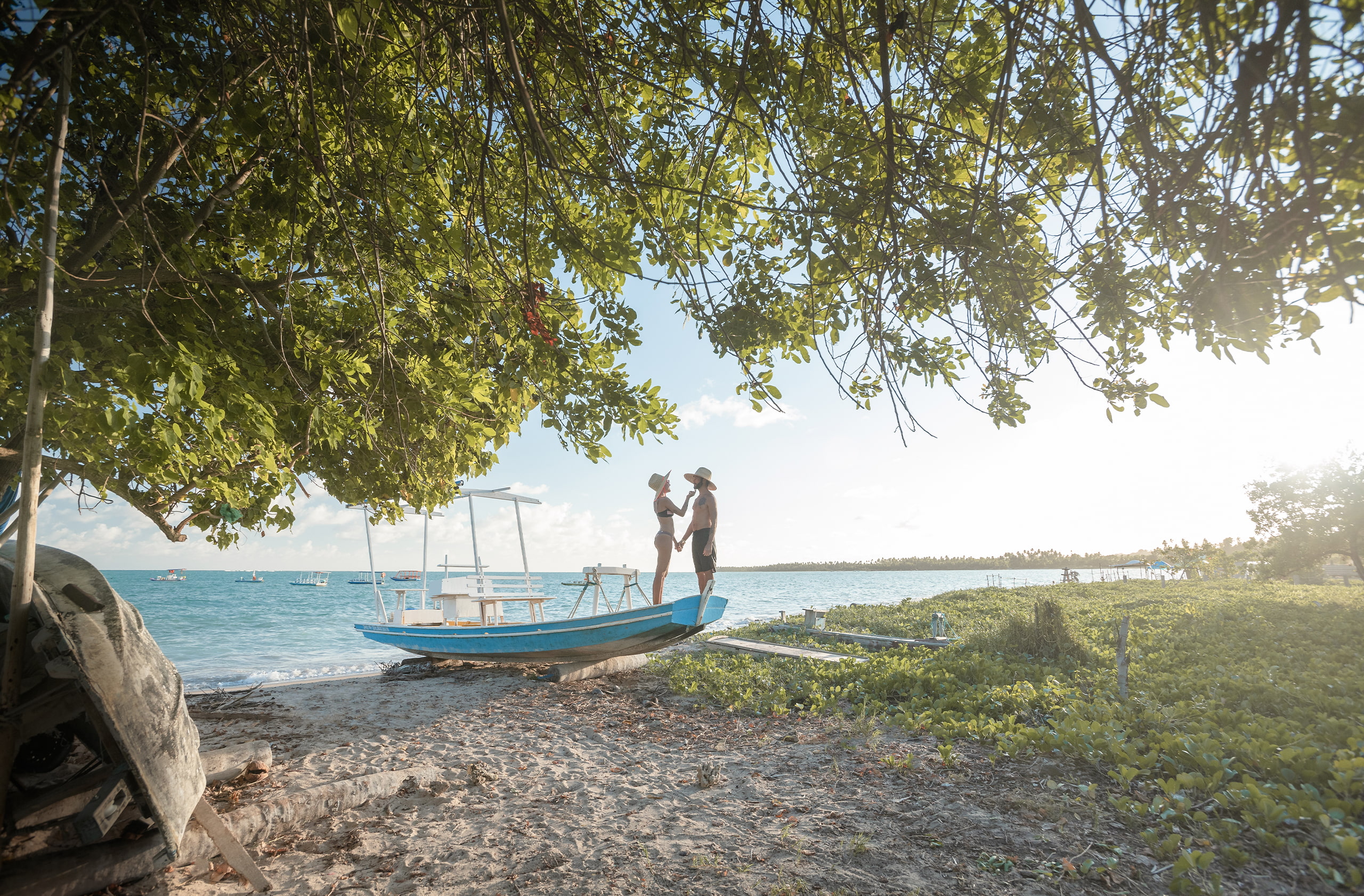 Casal em cima de um barco na areia da Praia de Patacho. Ao fundo há o mar de tom azul com alguns barcos, e a praia tem bastante vegetação.