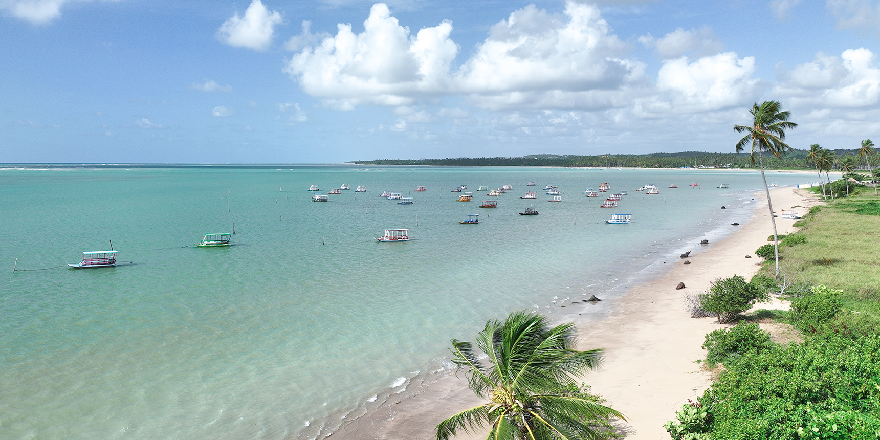 Vista aérea da Praia Patacho, com águas claras e calmas. Vários barcos pequenos estão ancorados na água, e a costa tem areia branca e muitas palmeiras.