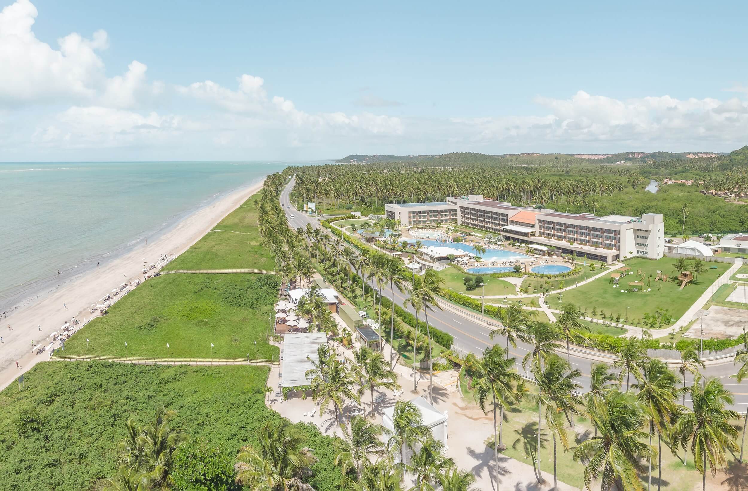 Vista aérea do Japaratinga Lounge Resort, um resort de praia no nordeste, em meio a muita vegetação e na beira-mar. É possível ver toda a sua estrutura, prédio e o complexo de piscinas, além de parte do mar.