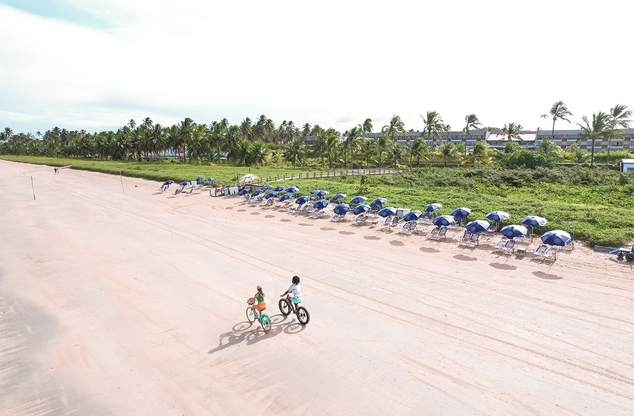 Um casal passeia de bicicleta na areia da praia em frente ao Japaratinga Lounge Resort, um resort de praia paradisíaco no litoral do Brasil. A vista conta com uma areia clara, guarda-sóis na areia e muita vegetação ao fundo.