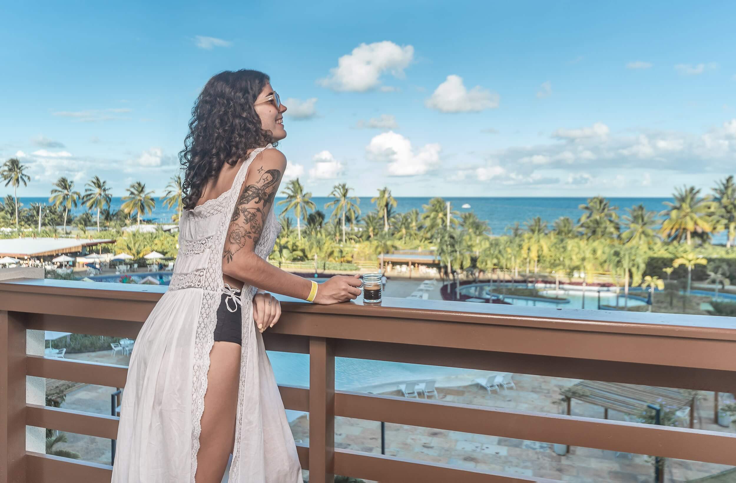 Uma mulher aprecia um momento tranquilo em uma sacada no Japaratinga Lounge Resort, segurando uma xícara de café. Ela contempla a deslumbrante vista das piscinas do resort e do mar azul que se estende até o horizonte.