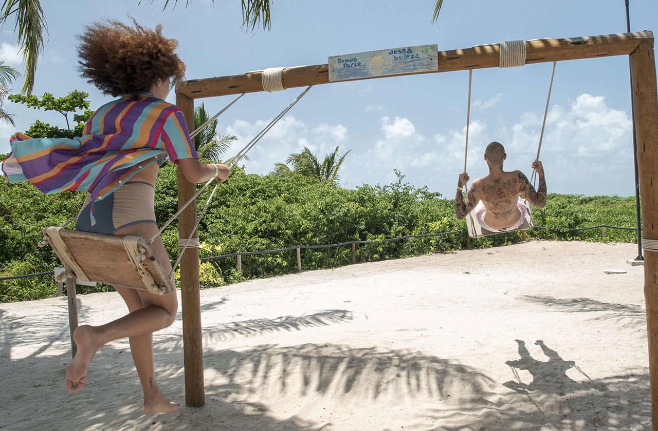 É possível ver uma casal utilizando balanços no Japaratinga Lounge resort. A estrutura está fincada na areia da praia e ao fundo vê-se vegetação e um dia ensolarado com nuvens brancas.