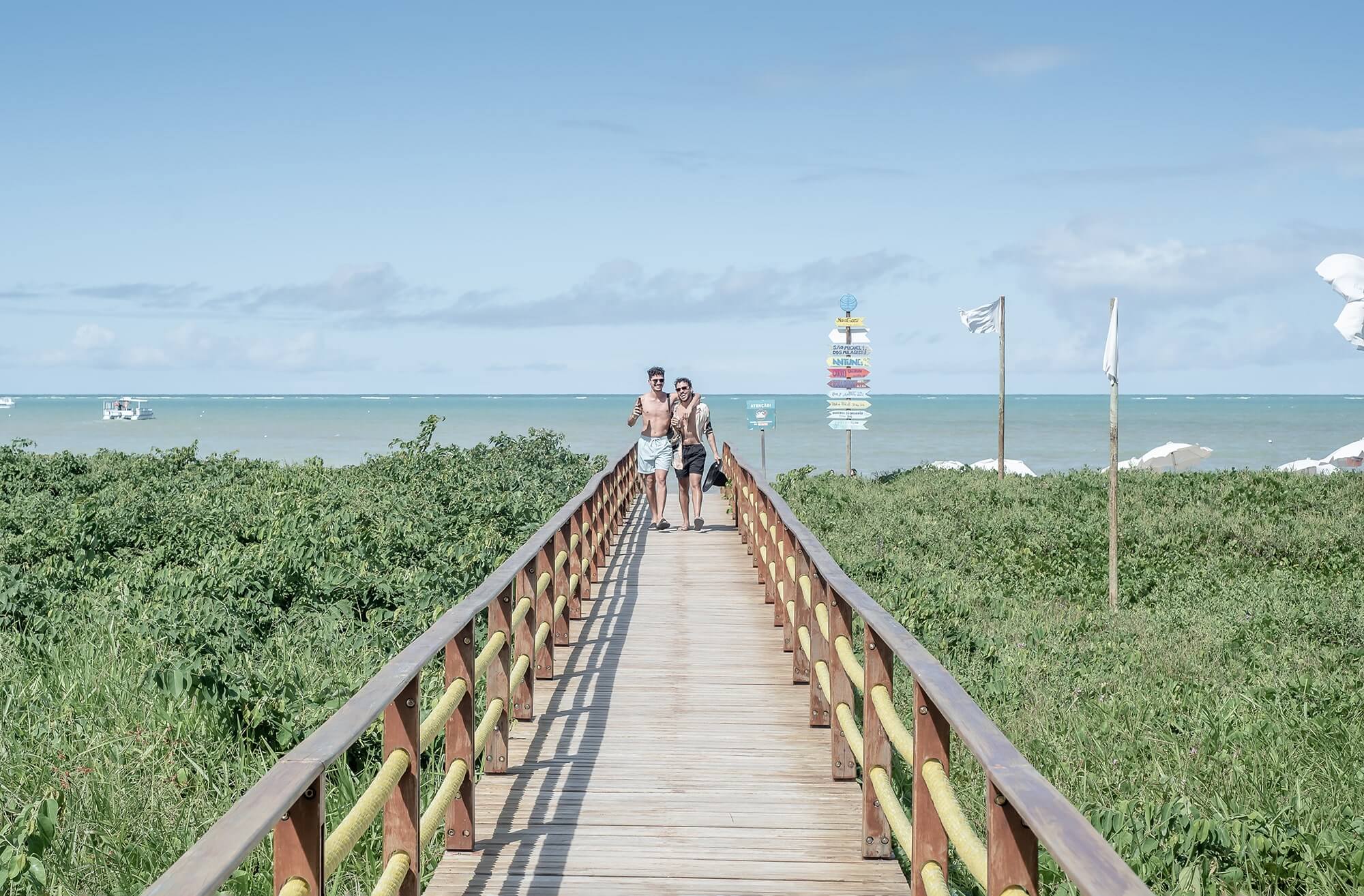 Um casal está caminhando na ponte que conecta a beira-mar de Japaratinga ao Resort. A paisagem ao redor é composta por uma vegetação e ao fundo vê-se a praia de Japaratinga e um céu azul com poucas nuvens.