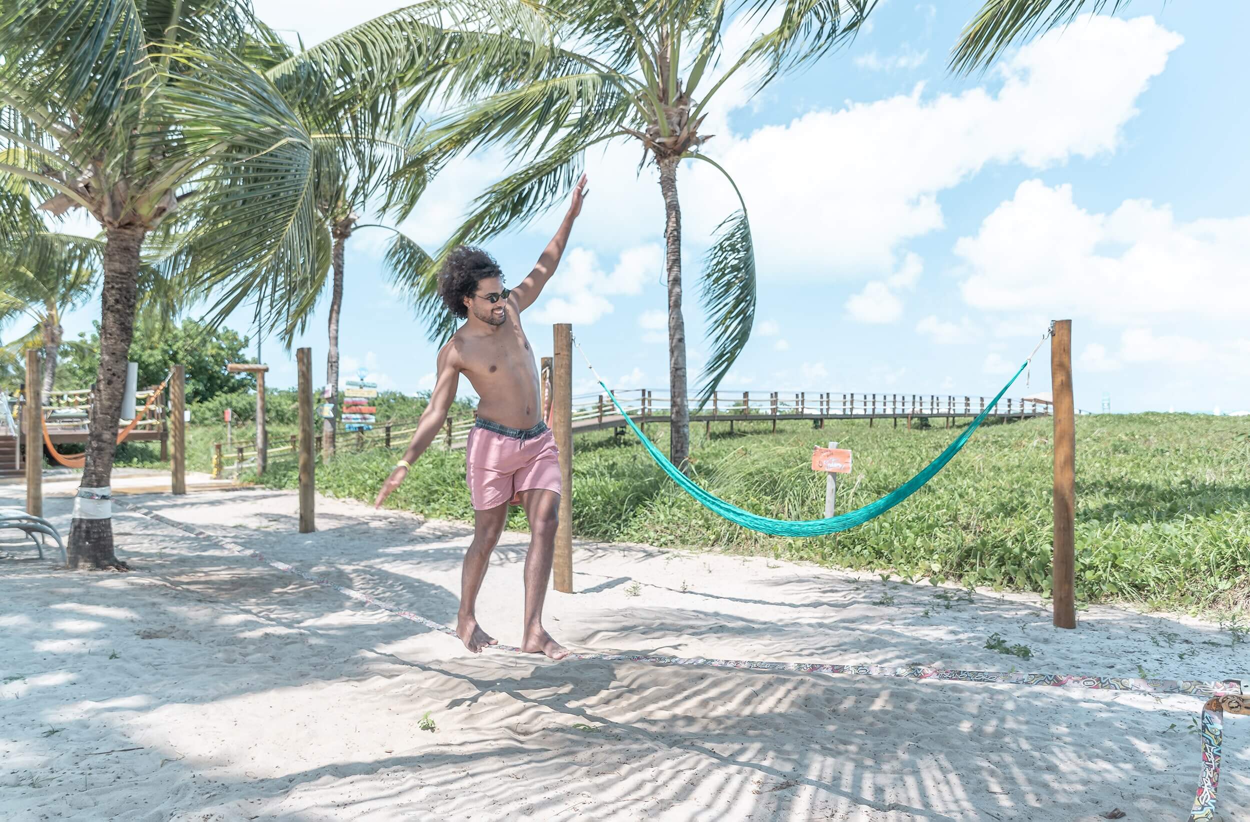 Um hóspede do Japaratinga Lounge Resort está se equilibrando em uma corda de slackline à beira-mar. Ela está presa entre árvores, acima de uma areia clara e ao fundo pode-se ver bastante vegetação e um céu azul.