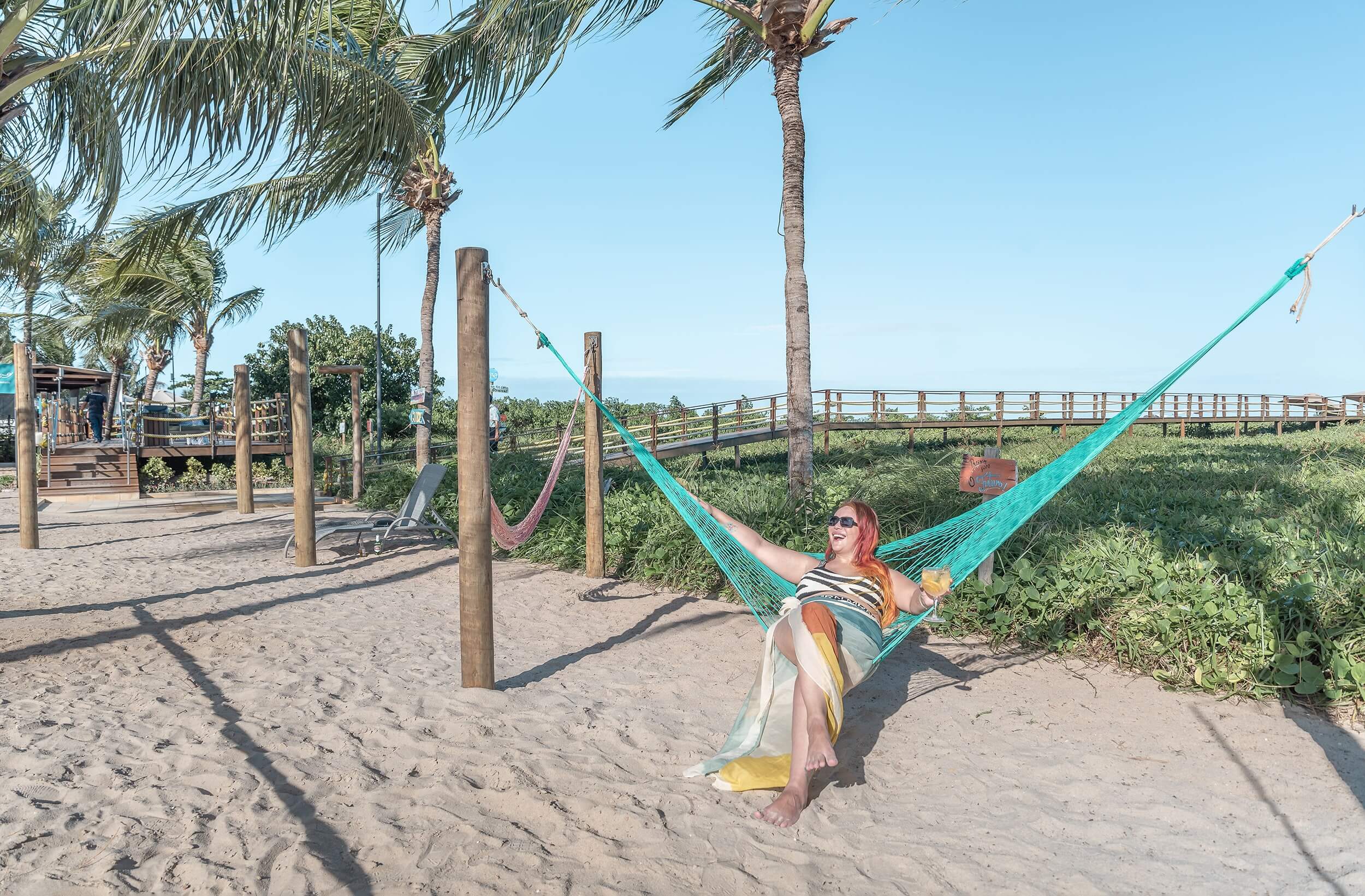 Uma mulher está deitada em uma rede verde amarrada em um estrutura de madeira redonda fincada na areia do Japaratinga Lounge Resort. Ao fundo pode-se ver vegetação, um céu azul e o horizonte marcado pelo mar.
