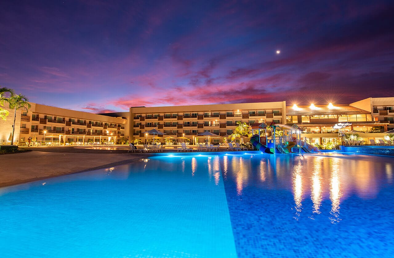 Uma cena noturna captura da piscina em tons azuis do Japaratinga Lounge Resort, onde as luzes amarelas do complexo de apartamentos ao fundo se refletem na água.