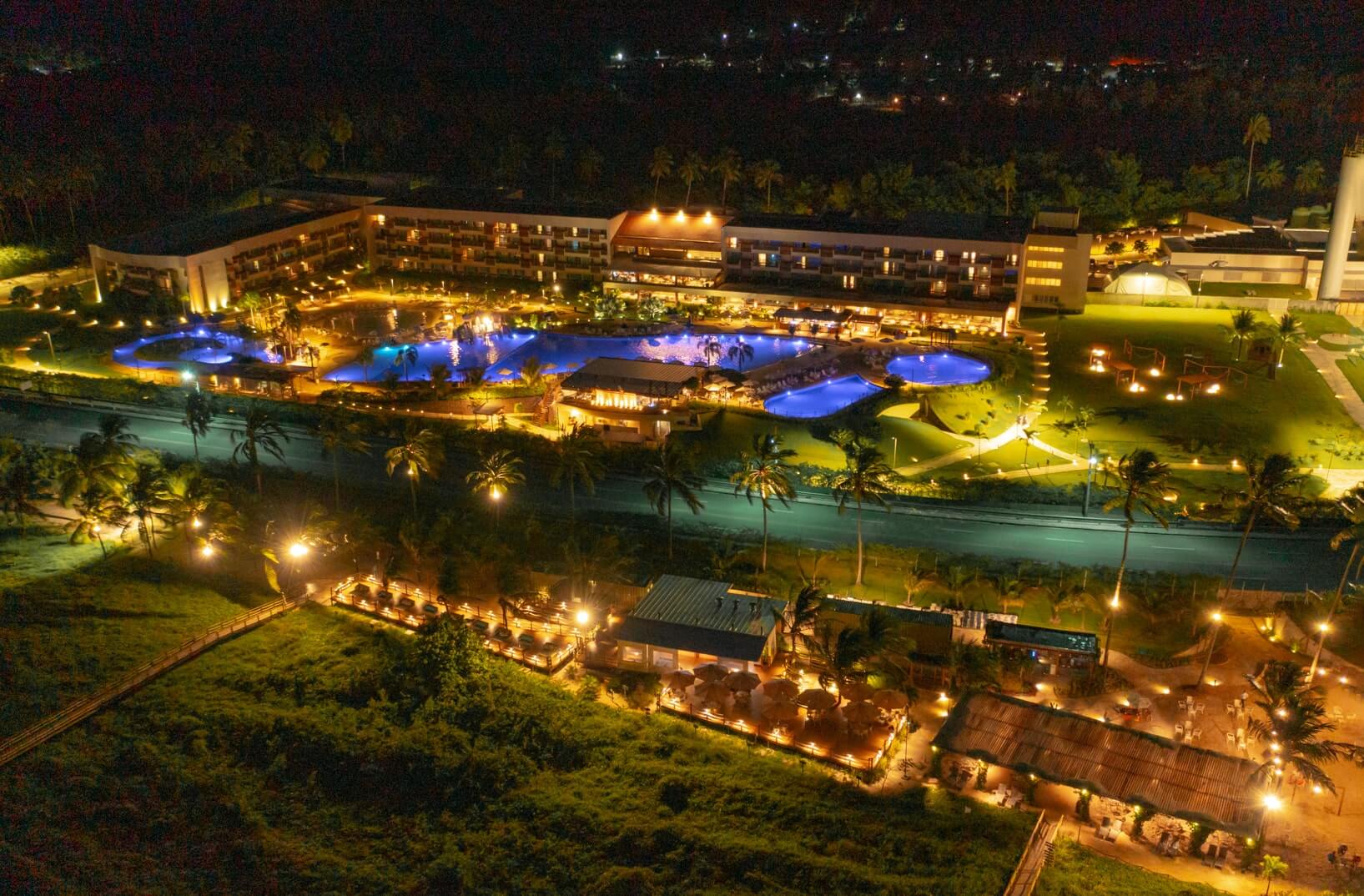 Vista aérea noturna da estrutura completa do Japaratinga Lounge Resort revela a magia das luzes que iluminam todoo resort, enquanto as piscinas exibem reflexos em tons de azul.