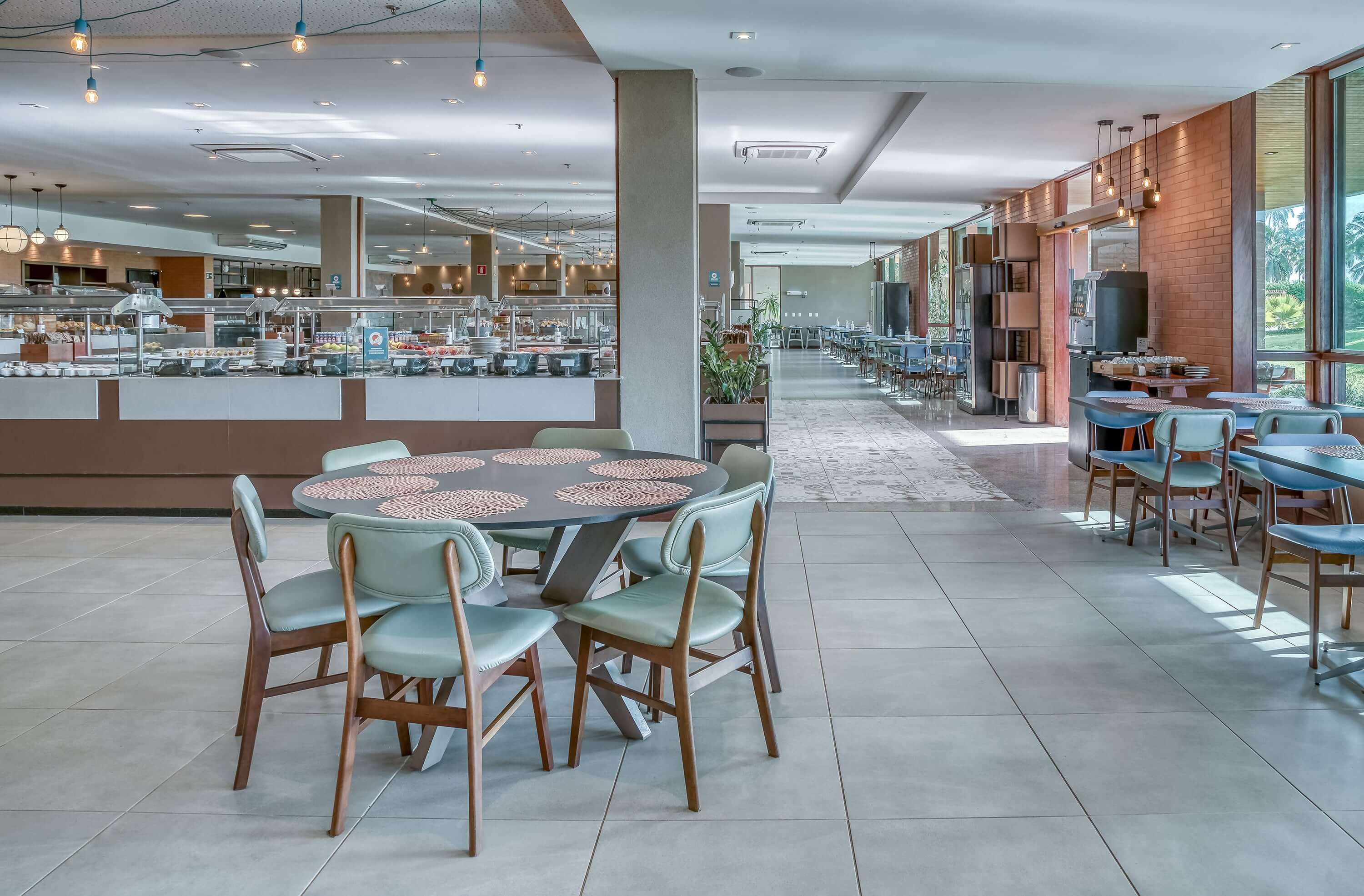 A imagem captura a atmosfera acolhedora do Restaurante Tabôa no Japaratinga Lounge Resort. Mesas e cadeiras em tons azuis e marrons e ao fundo vê-se parte das estações de self-service.