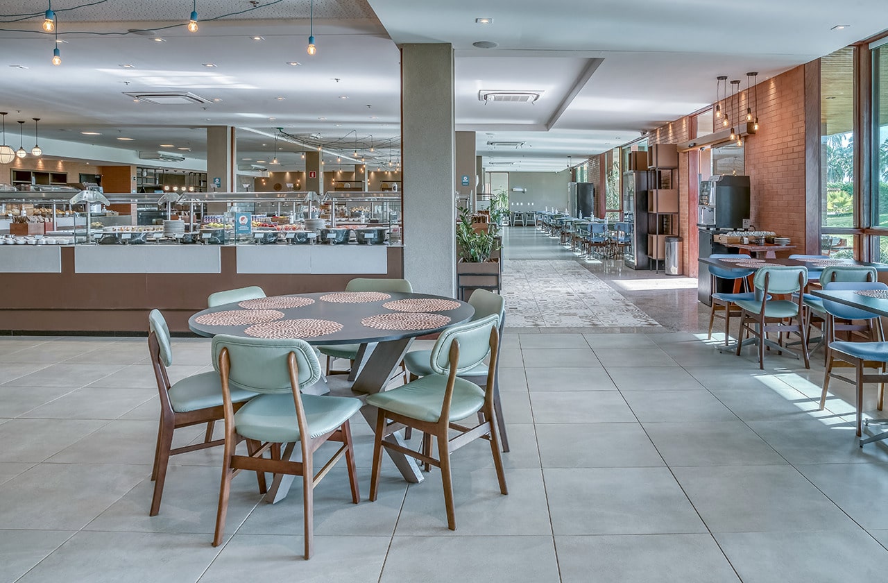 É possível ver uma casal utilizando balanços no Japaratinga Lounge resort. A estrutura está fincada na areia da praia e ao fundo vê-se vegetação e um dia ensolarado com nuvens brancas.