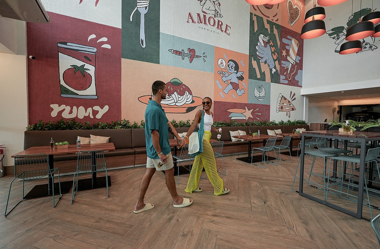 Um casal está caminhando na ponte que conecta a beira-mar de Japaratinga ao Resort. A paisagem ao redor é composta por uma vegetação e ao fundo vê-se a praia de Japaratinga e um céu azul com poucas nuvens.