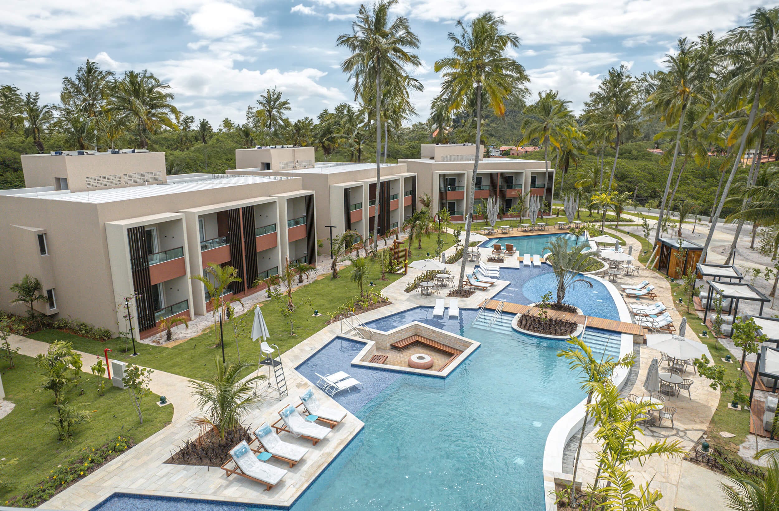 A imagem mostra uma vista aérea do complexo de resort. Há um conjunto de edifícios com arquitetura moderna, pintados em cores neutras com detalhes em terracota, alinhados em torno de uma área de piscina central. A piscina possui várias seções, incluindo uma área rasa para espreguiçadeiras submersas e um jacuzzi. Espreguiçadeiras e guarda-sóis estão dispostos ao redor da piscina, convidando ao relaxamento e ao lazer.