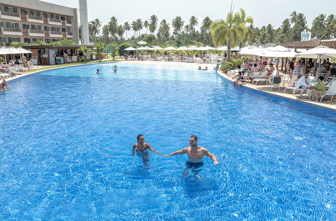 Um casal passeia de bicicleta na areia da praia em frente ao Japaratinga Lounge Resort, um resort de praia paradisíaco no litoral do Brasil. A vista conta com uma areia clara, guarda-sóis na areia e muita vegetação ao fundo.