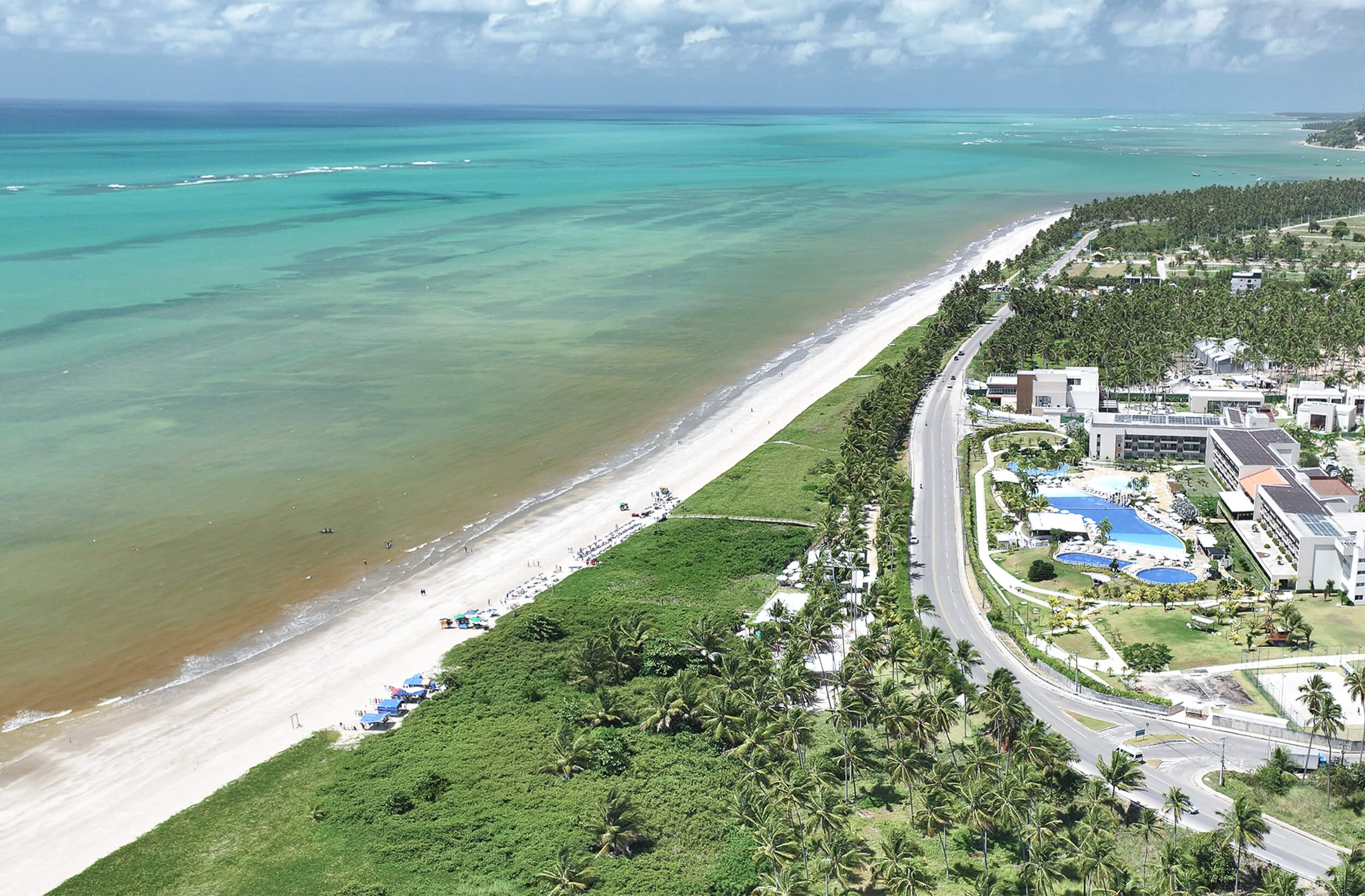 A imagem oferece uma vista aérea da praia de Japaratinga. A praia de areia branca se estende ao longo de uma água azul-turquesa, onde é possível ver variações de cor indicando mudanças na profundidade do mar. Uma estrada pavimentada serpenteia paralelamente à costa, ladeada por uma fileira de coqueiros, ao lado está construções do resort.