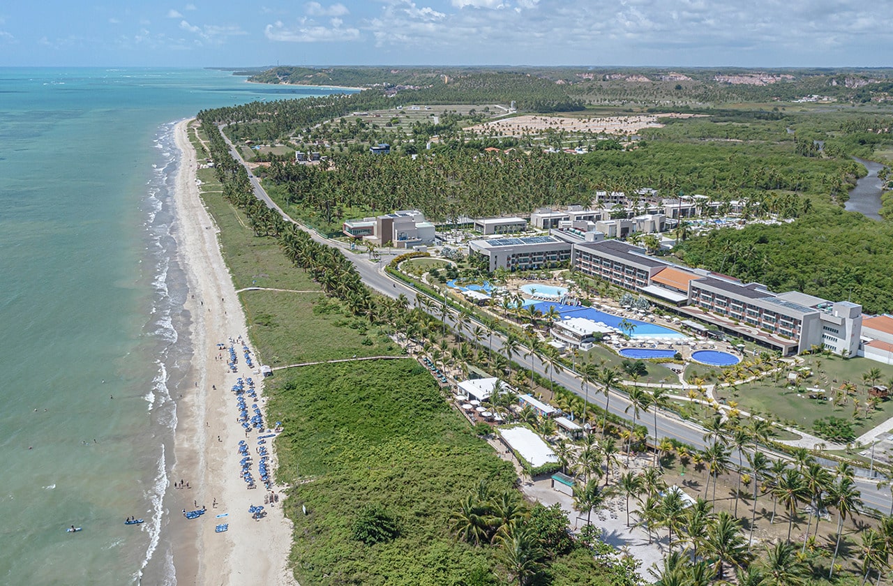 A imagem oferece uma vista aérea da praia de Japaratinga. A praia de areia branca se estende ao longo de uma água azul-turquesa, onde é possível ver variações de cor indicando mudanças na profundidade do mar. Uma estrada pavimentada serpenteia paralelamente à costa, ladeada por uma fileira de coqueiros, ao lado está construções do resort.