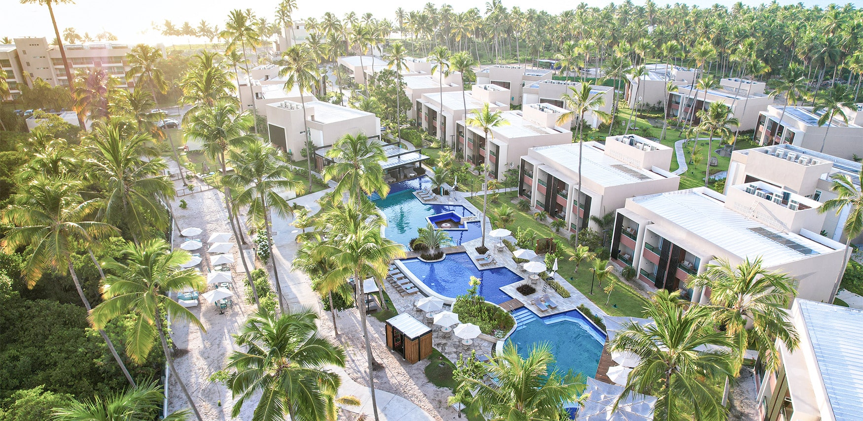 Vista aérea do Japaratinga Lounge Resort com coqueiros altos, piscinas azuis e bangalôs modernos, transmitindo uma energia de férias de verão e relax.