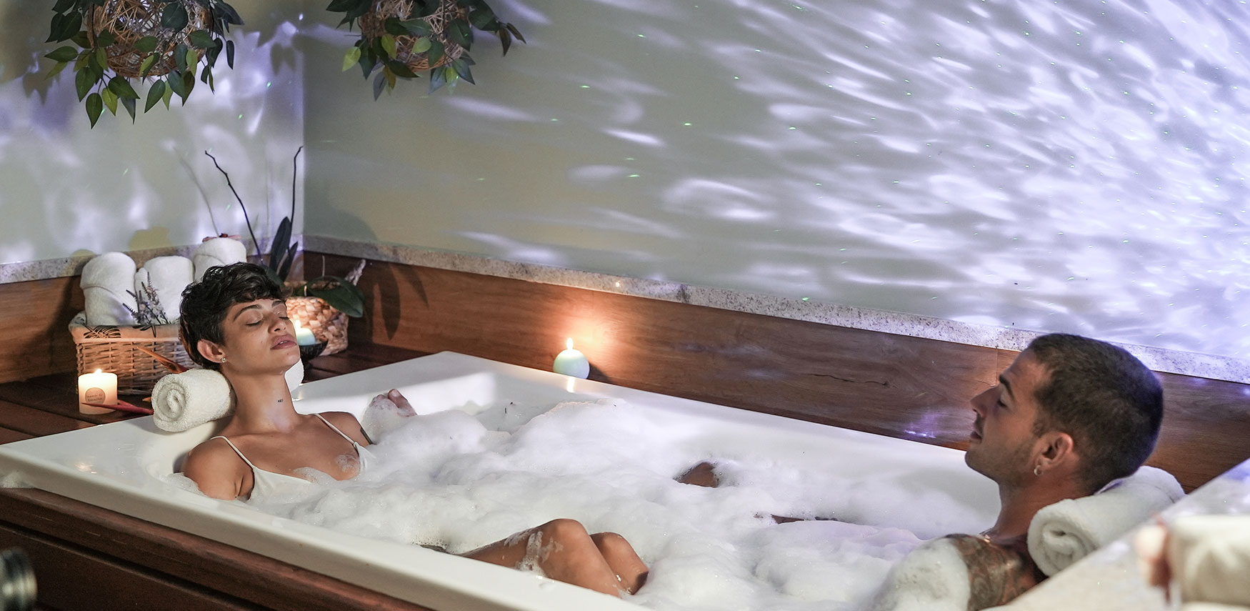 Casal relaxando juntos em uma banheira de hidromassagem em um ambiente de spa. A iluminação suave, com plantas, velas acesas e a espuma criam uma atmosfera de calma e relaxamento.