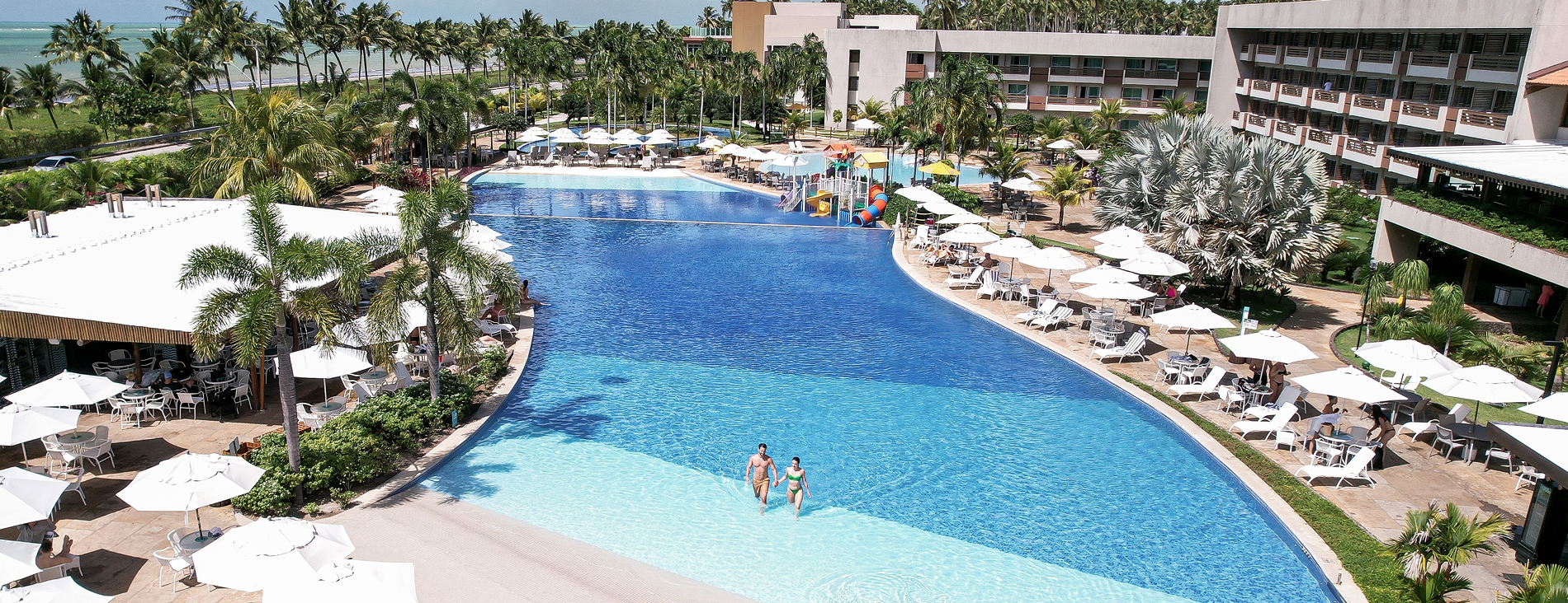 Vista aérea da piscina principal. O entorno é repleto de guarda-sóis e mesas brancas, coqueiros e o edifício principal do resort. Ao fundo, o mar azul com a linha de coqueiros na praia sob um céu parcialmente nublado.