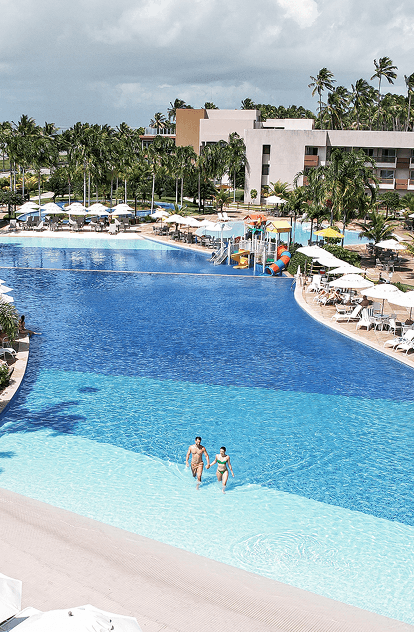 black friday japaratinga lounge resort Vista aérea da piscina principal. O entorno é repleto de guarda-sóis e mesas brancas, coqueiros e o edifício principal do resort. Ao fundo, o mar azul com a linha de coqueiros na praia sob um céu parcialmente nublado.