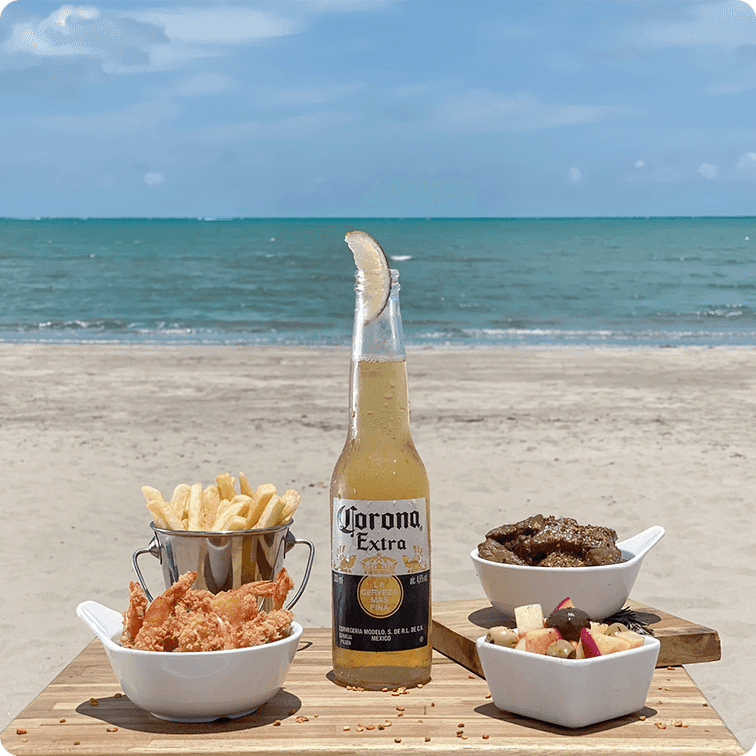 Uma garrafa de cerveja Corona Extra com uma fatia de limão, rodeada por petiscos em bowls brancos com batata frita, camarão empanado e carne, além de frutas. Tudo está disposto sobre uma tábua de madeira na areia da praia, com o mar e o horizonte ao fundo.