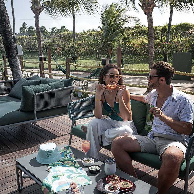Casal sorrindo e relaxando em confortáveis sofás de área externa no deck de madeira de o Japaratinga Lounge Resort. Eles estão compartilhando petiscos e bebidas, com a mulher segurando um pedaço de pizza. A cena é sombreada por coqueiros e rodeada por vegetação tropical.