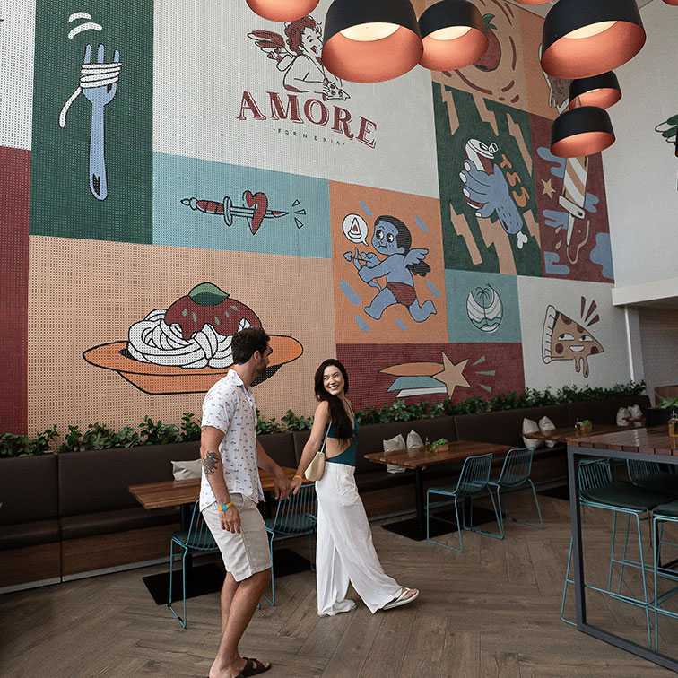 Casal jovem sorrindo e de mãos dadas, caminhando em direção à mesa da Amore Forneria. A parede de fundo é um grande mural de azulejos coloridos com ilustrações de comida e a palavra "AMORE". O ambiente tem iluminação baixa e luminárias pendentes.